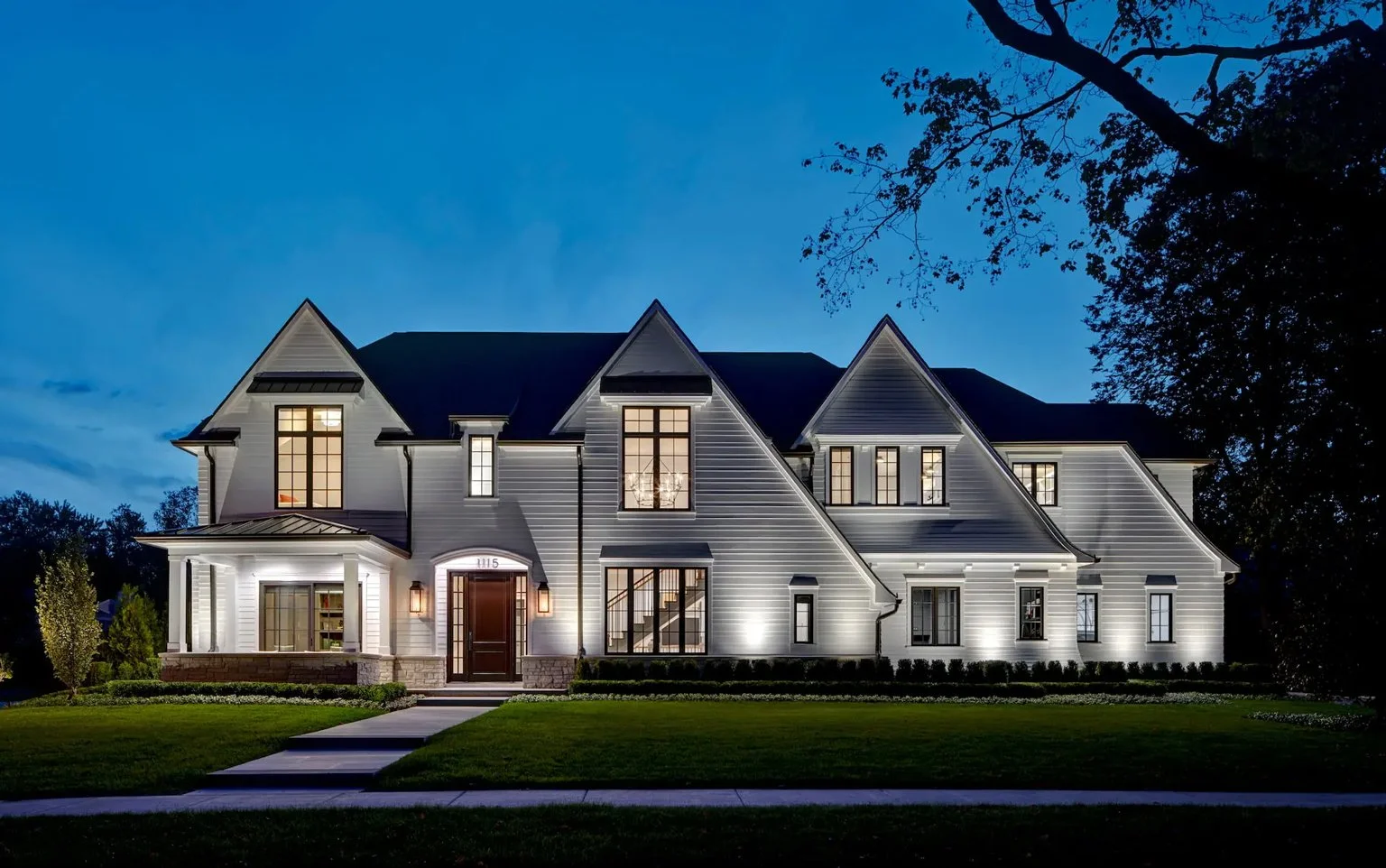 A modern, white, two-story house with multiple gabled roofs and large windows, illuminated at night with exterior lighting, surrounded by a manicured lawn and trees.