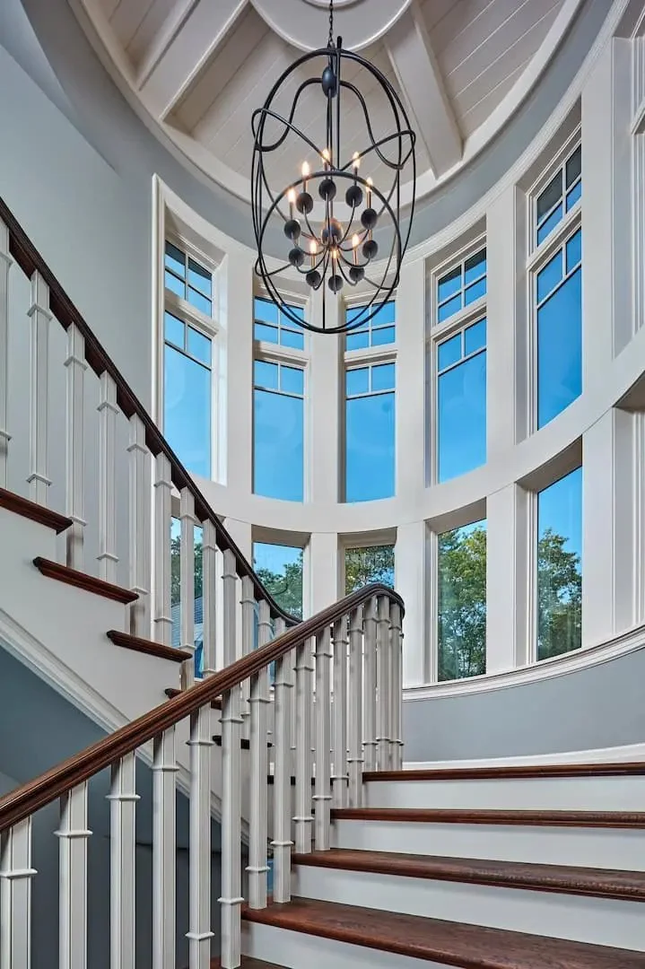 Interior view of a curved staircase with white banisters and dark wooden handrails, leading up to a large, round windowed wall with multiple windows and a modern chandelier hanging from the ceiling.