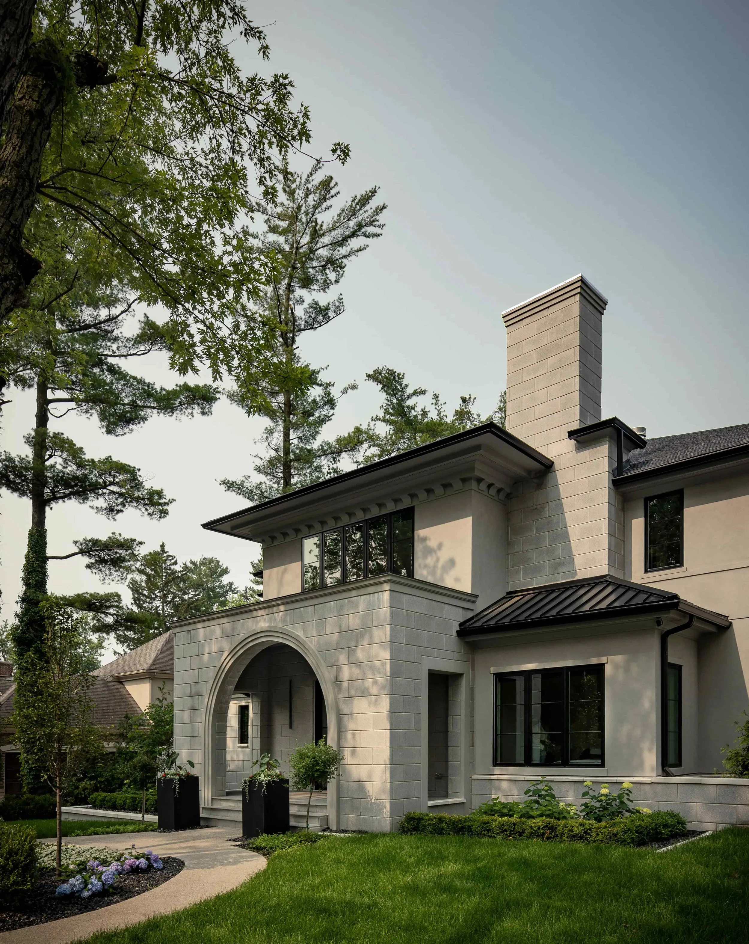Modern two-story house with a stone facade and black window frames, surrounded by green lawn and tall trees.