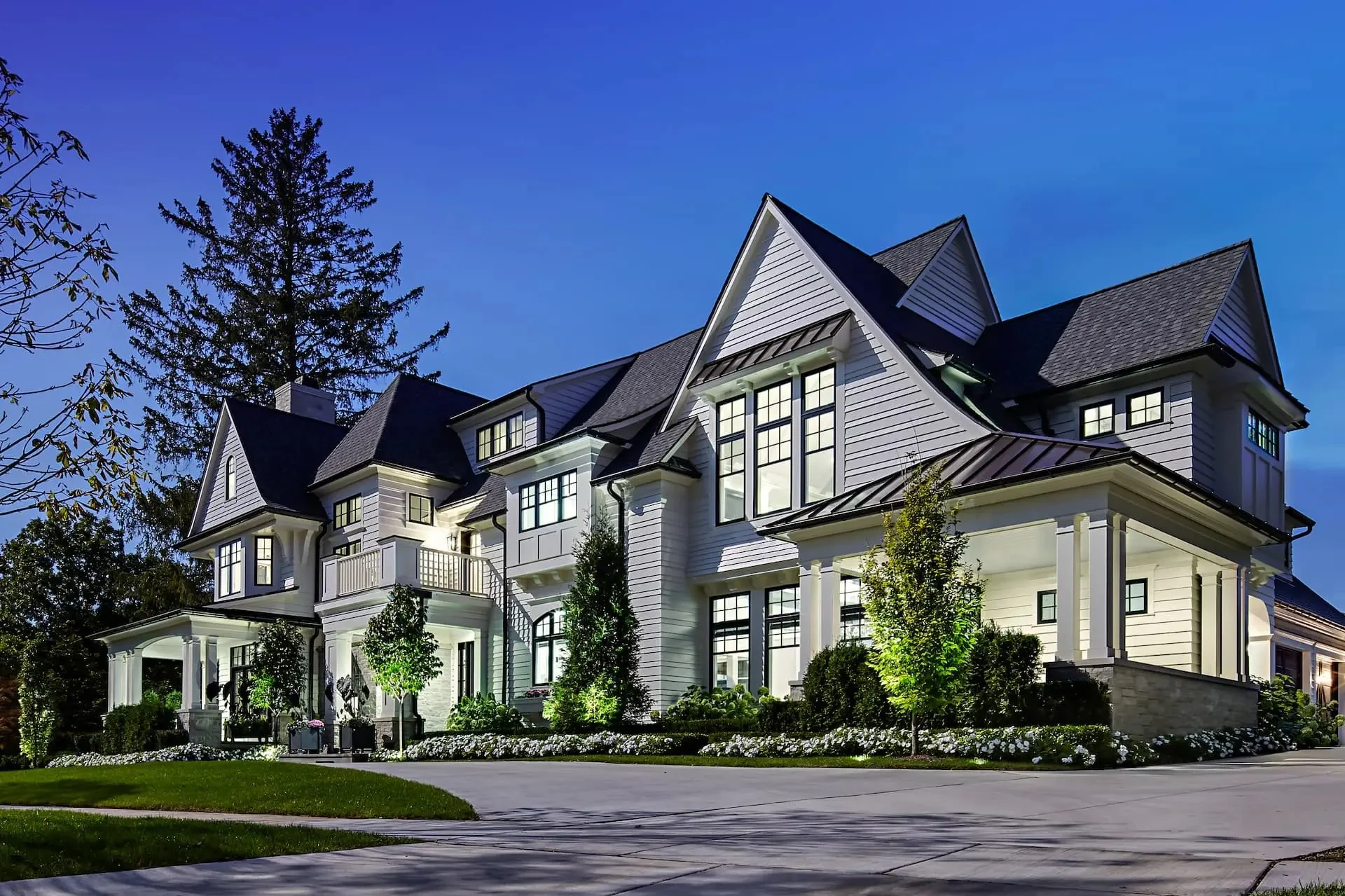 Large white mansion with multiple gabled roofs and large windows, illuminated at night, surrounded by landscaped garden.