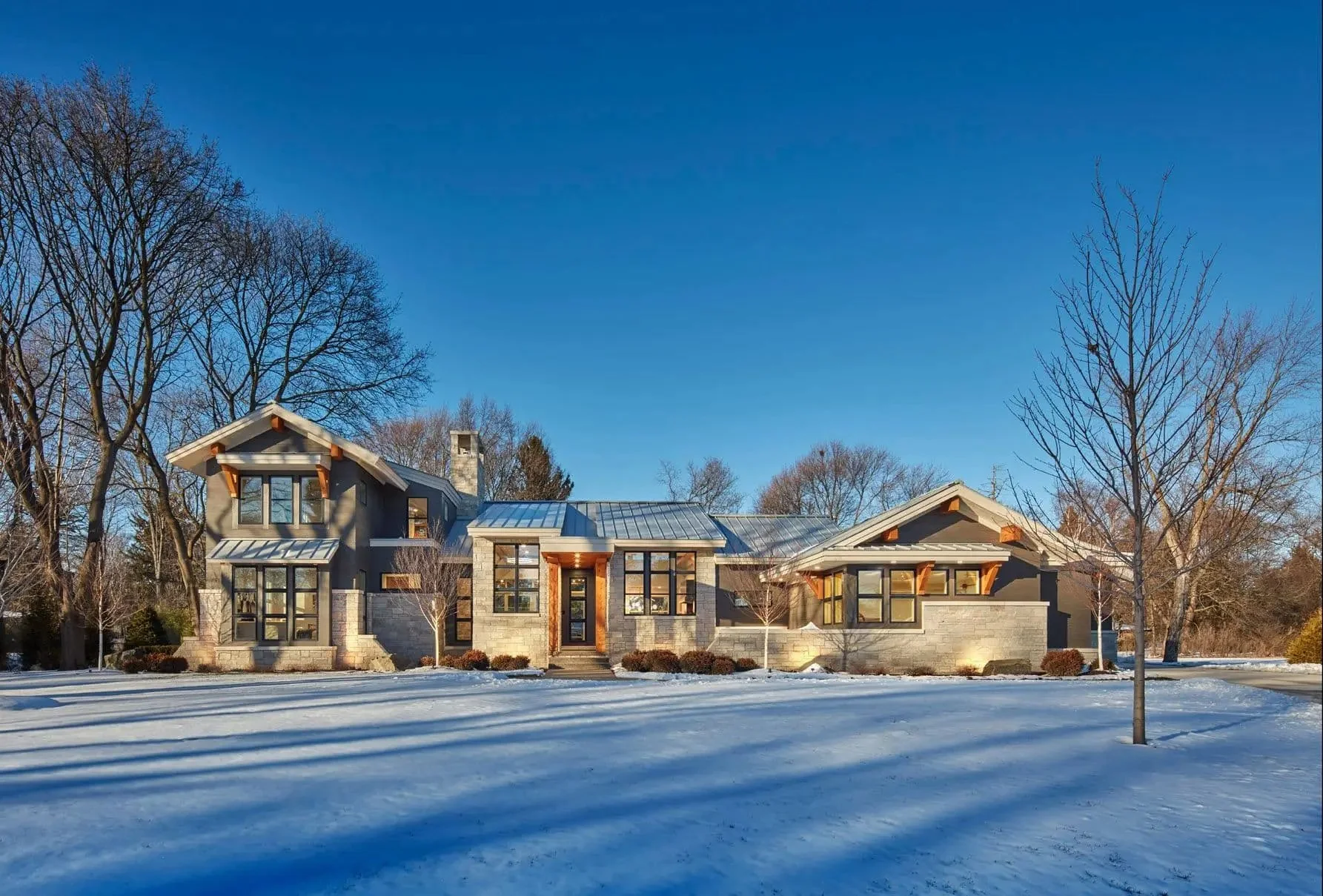 Modern house with stone and wood exterior, large windows, metal roof, surrounded by leafless trees and snow on the ground, under a clear blue sky.
