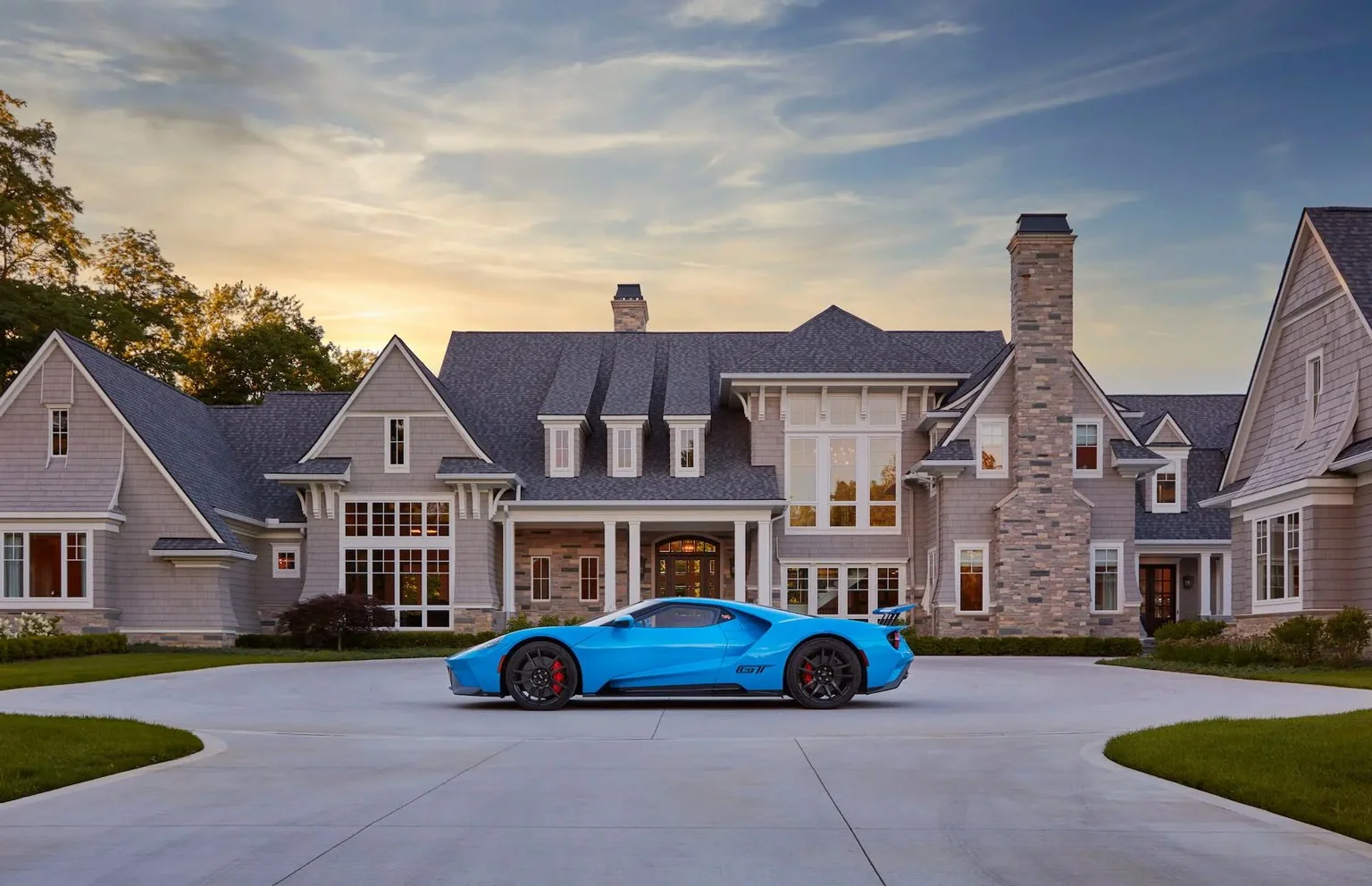 A blue sports car parked in front of a large, upscale house during sunset.