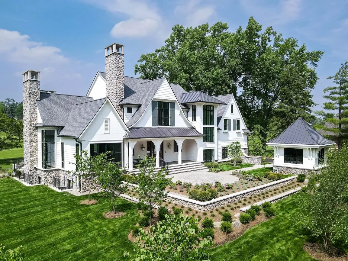 Large white house with multiple steep gabled roofs, stone chimneys, and a separate small pavilion, surrounded by a well-maintained lawn and garden with trees and shrubs, under a blue sky.
