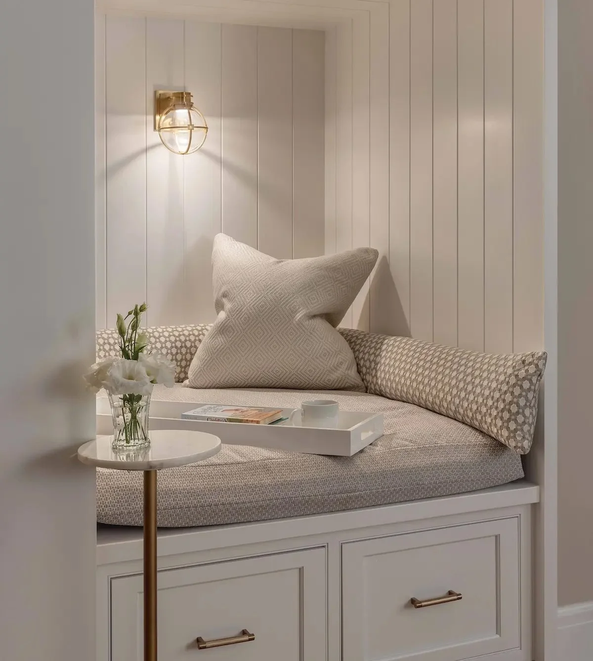 A cozy, built-in window seat with cushions, a small side table with flowers, a book, and a cup, set against white paneled walls with a brass wall sconce lighting.