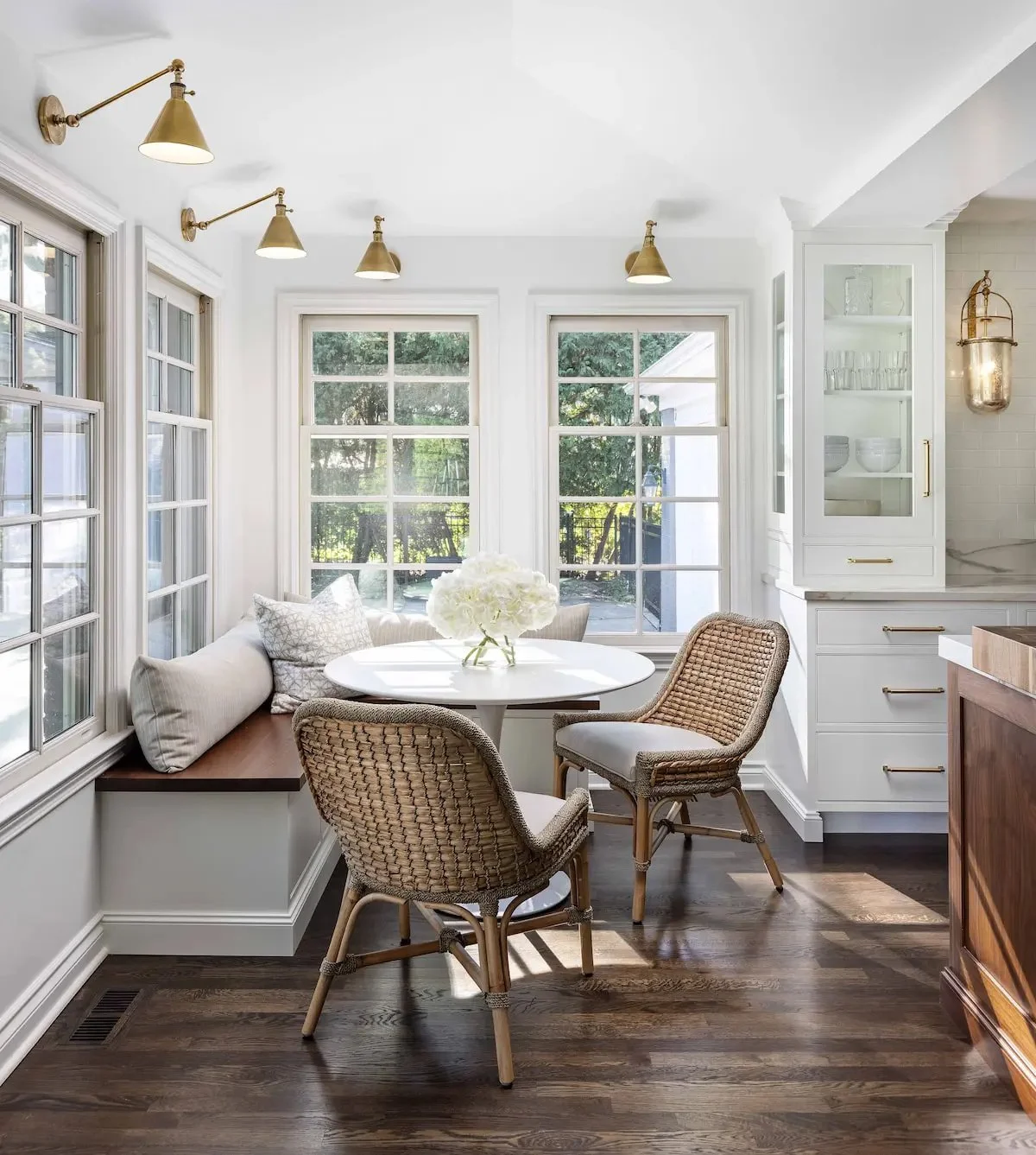 Bright dining nook with white walls, large windows, a white round table with a flower arrangement, wicker and upholstered chairs, and a built-in bench with pillows. Gold wall sconces and a tall white cabinet are visible.