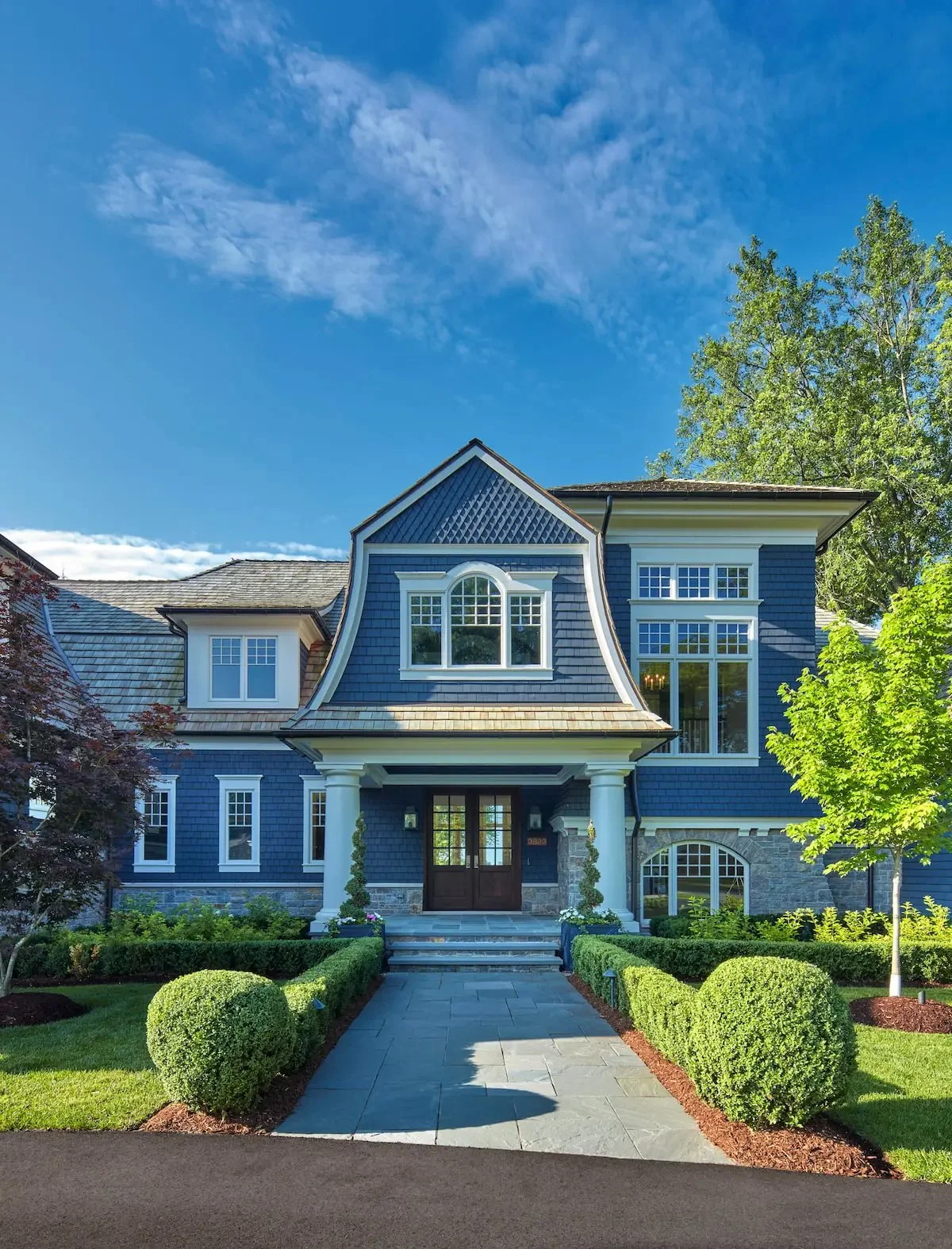 Front view of a blue house with multiple large windows, white columns, and a well-kept lawn with trimmed bushes and trees.