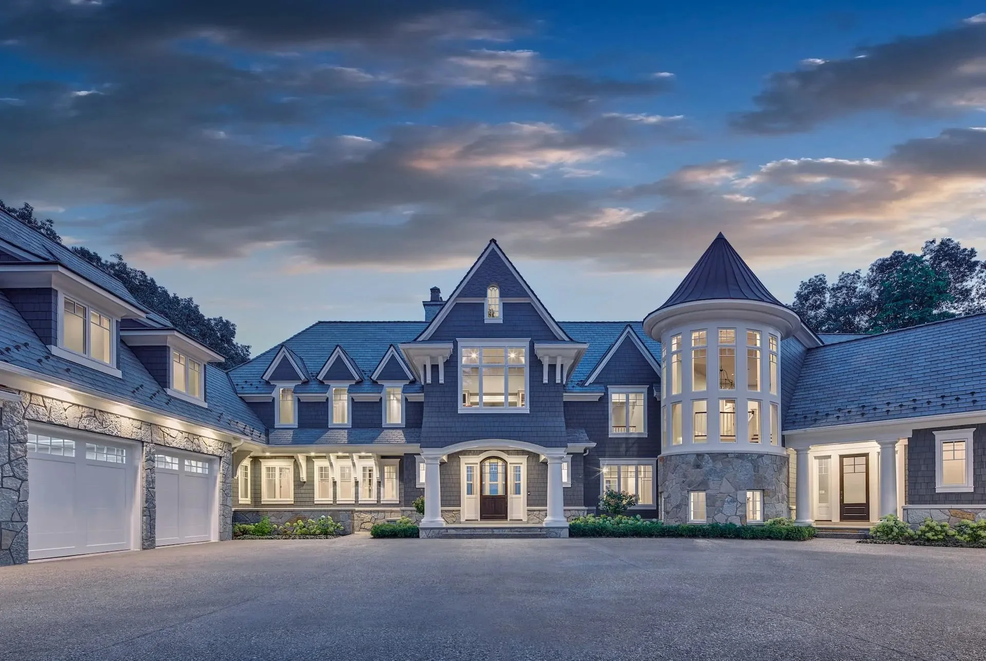 Large luxury house with multiple gabled roofs, a tower, and a three-car garage, illuminated at dusk.