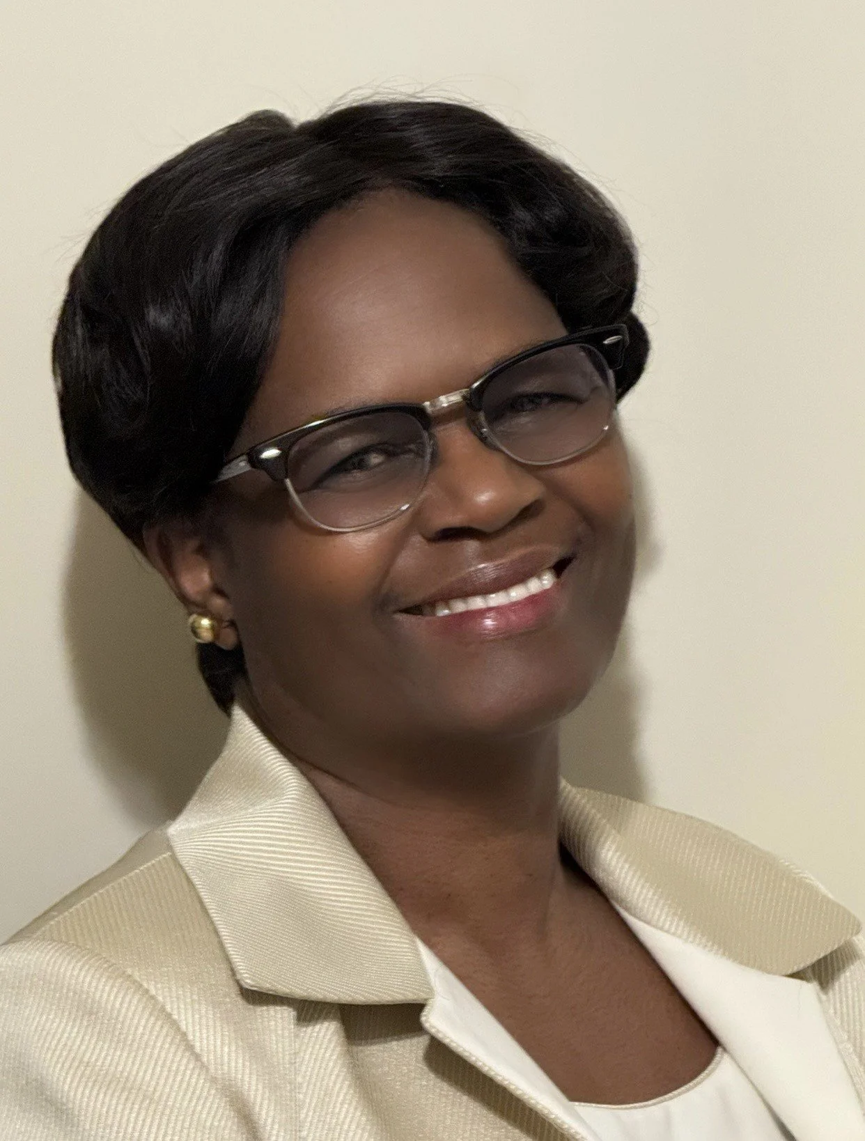A smiling woman with short black hair, glasses, and earrings, wearing a beige business suit, posing against a plain light-colored background.