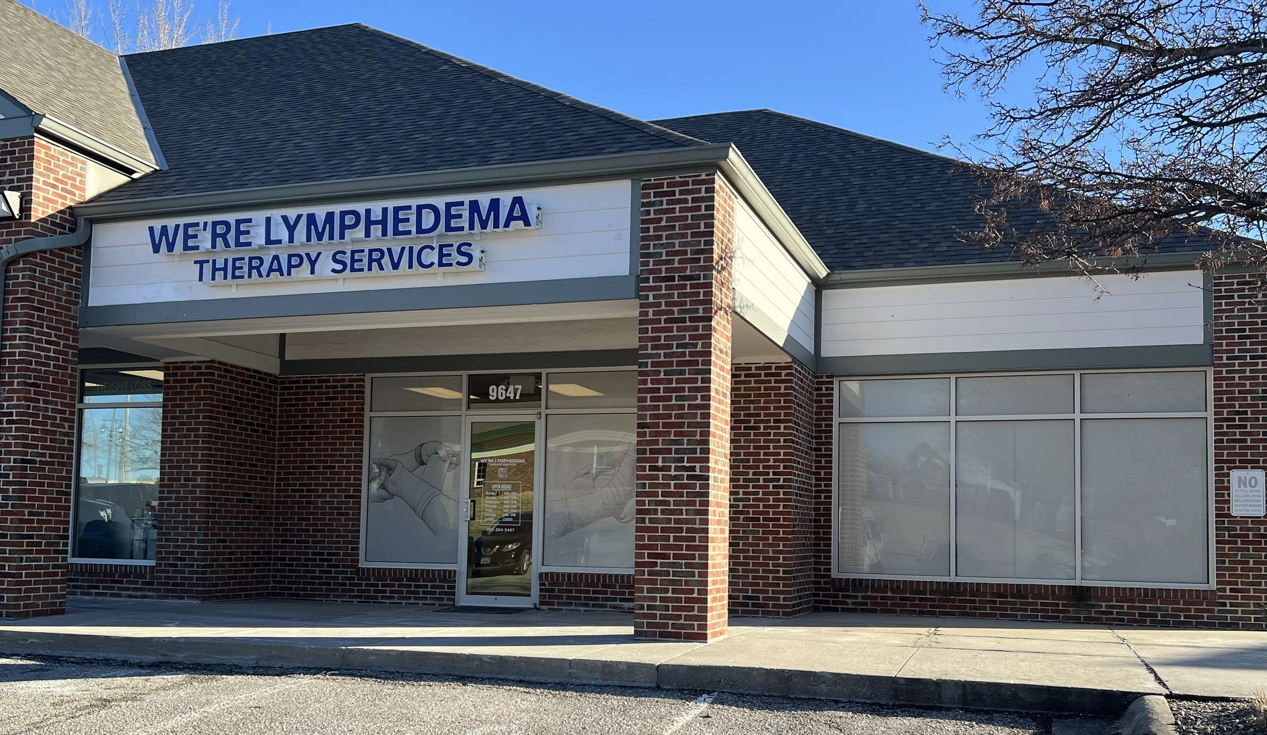 Brick building with a sign that reads "WE'RE LYMPHEDEMA THERAPY SERVICES" above the entrance. The building has a glass door and windows, and appears to be a healthcare or therapy clinic.