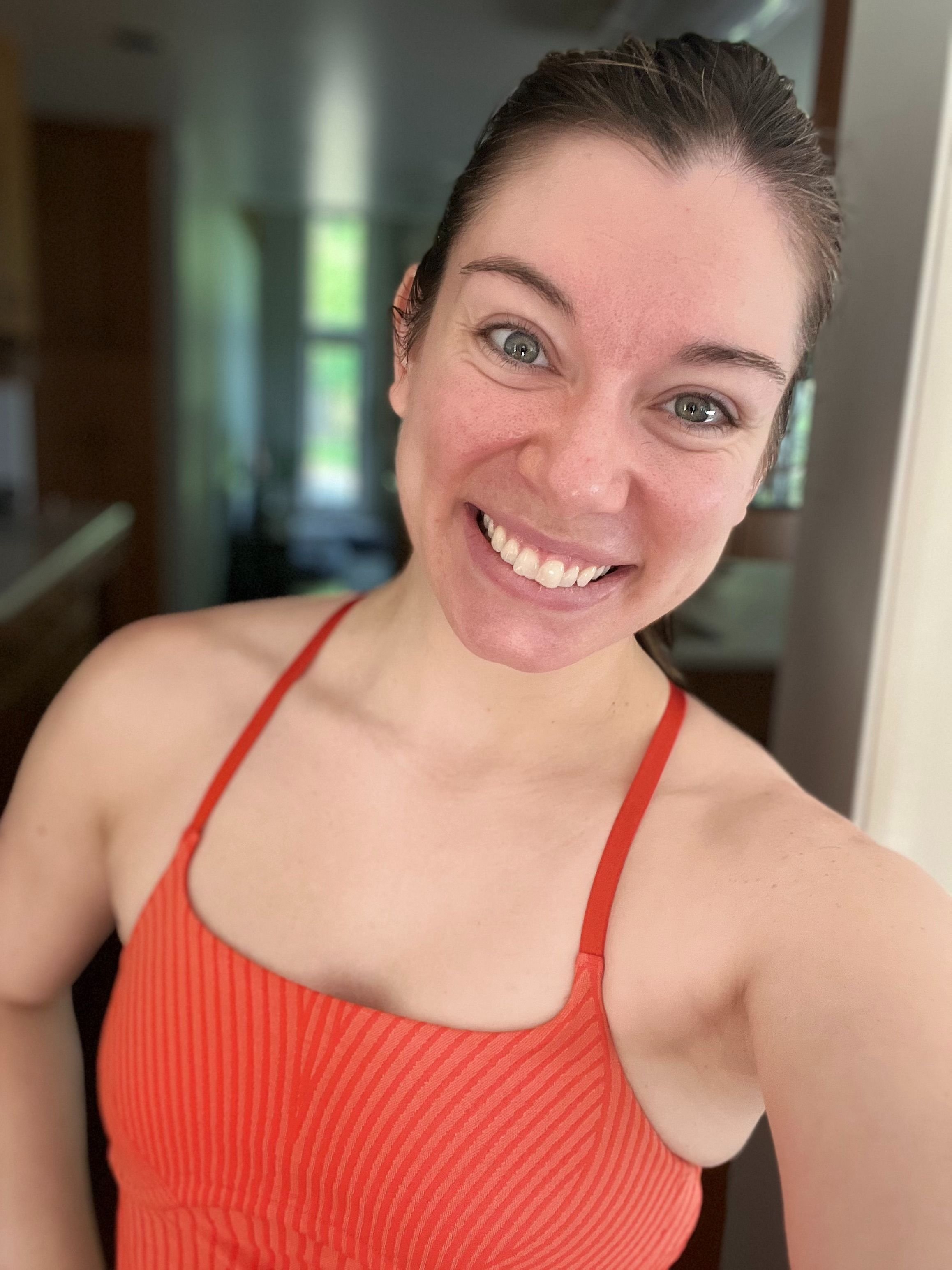 A woman with wet hair smiling at the camera, wearing a red striped tank top, indoors with a blurred background showing windows and furniture.
