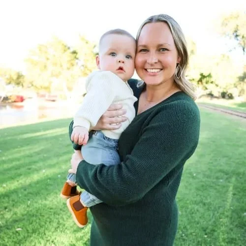 A woman smiling and holding a baby outdoors on a grassy area with trees in the background.