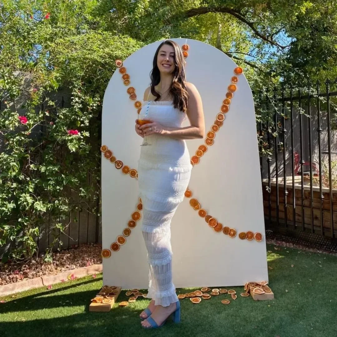 A woman in a white dress holding a drink, standing outdoors in front of a large white board decorated with orange slices, with greenery and trees in the background.