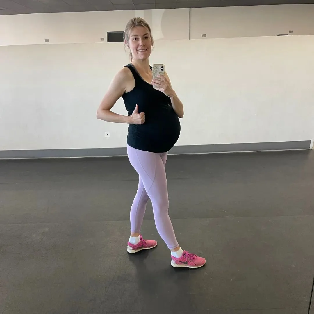 Pregnant woman taking a selfie in a gym mirror, smiling, wearing a black tank top, light pink leggings, and pink sneakers.
