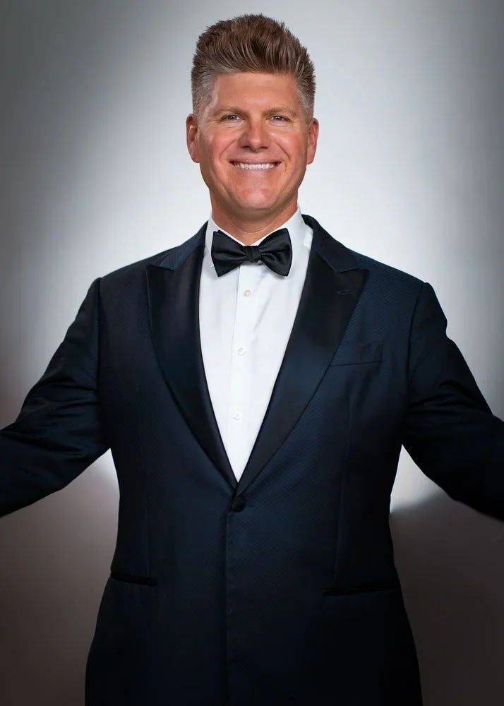 A man in a tuxedo with a bow tie smiling at the camera against a gray background.