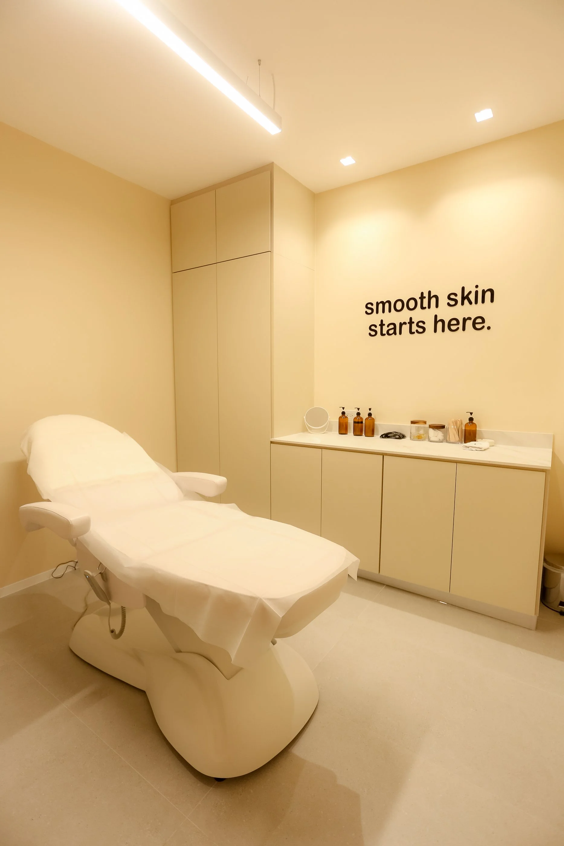 A clean, minimalist spa or skincare treatment room with a white treatment bed, beige walls, and a counter with bottles and skincare products. The wall features the phrase 'smooth skin starts here.'