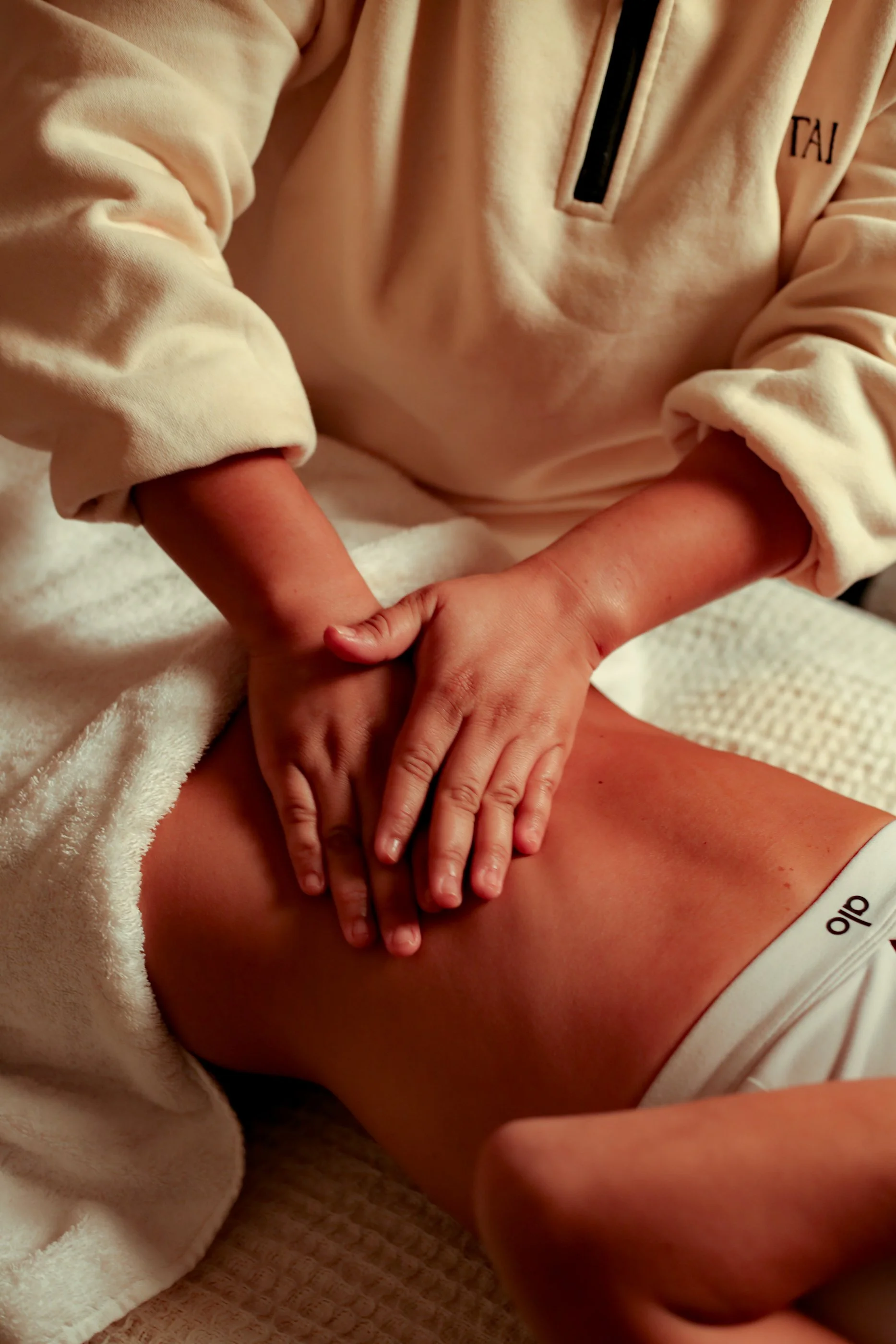 A person giving a massage to another person's back and thigh, with both individuals lying on a textured white surface. The person receiving the massage is wearing white underwear, and the massage is focused on the lower back and upper thigh area.