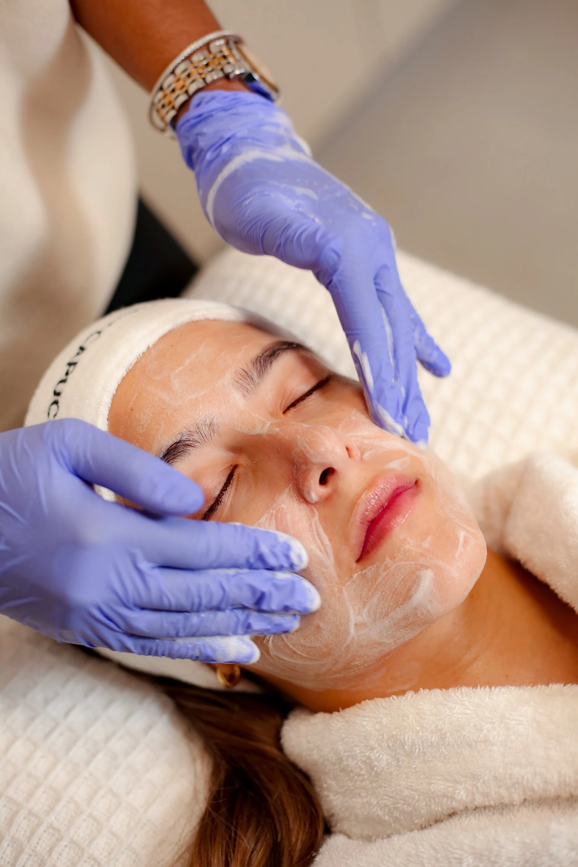 A woman receives a facial treatment, lying down with her eyes closed, wearing a towel and headband, while a skincare professional applies a white facial mask with gloved hands.