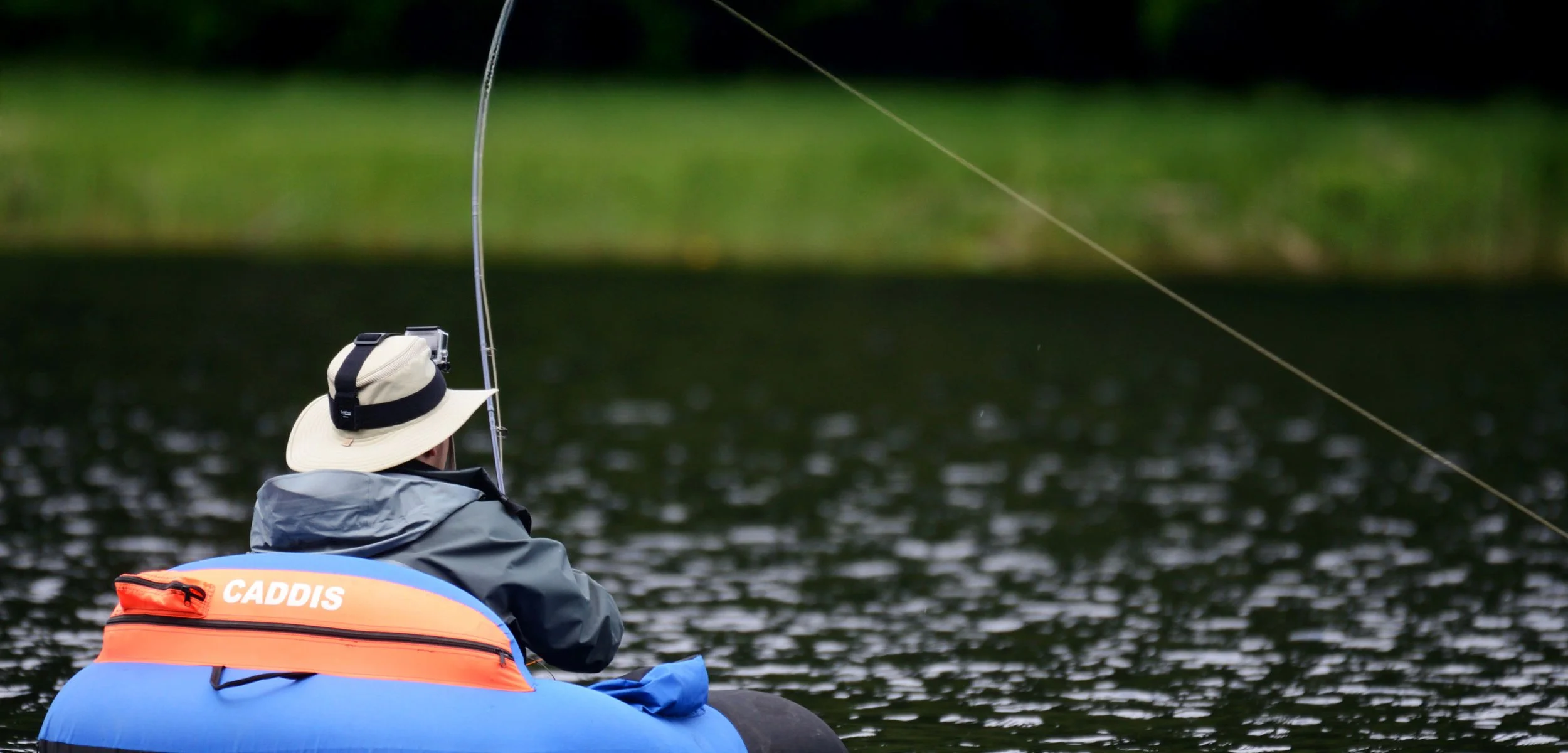 Person in a wide-brimmed hat and rain jacket fishing from a canoe on a calm body of water.