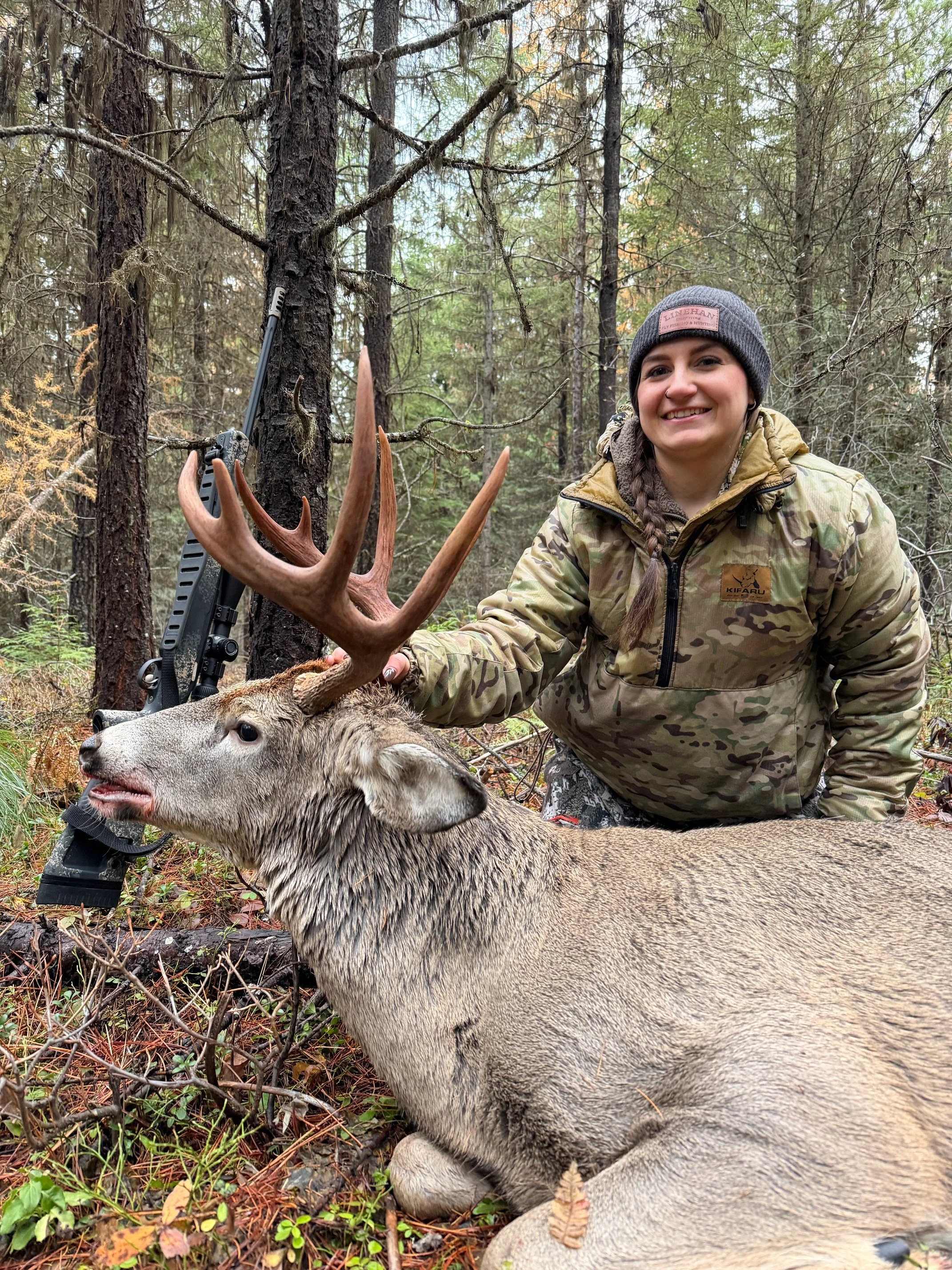 woman in camo clothing with rifle and whitetail buck in wooded area of Yaak Valley, Montana.