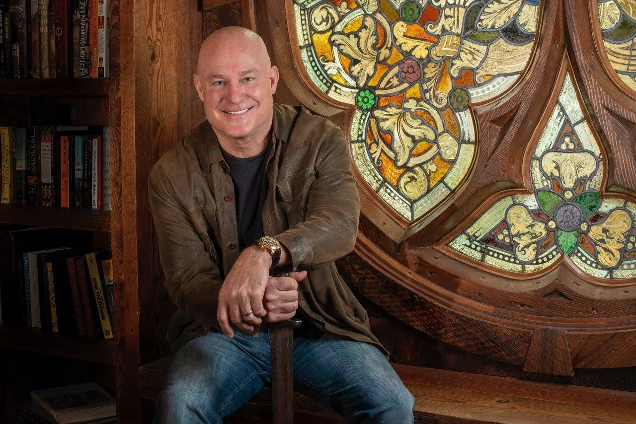 A smiling man with a shaved head, wearing a brown leather jacket, black shirt, and jeans, sitting in front of a stained glass window and a bookshelf in a wooden room.
