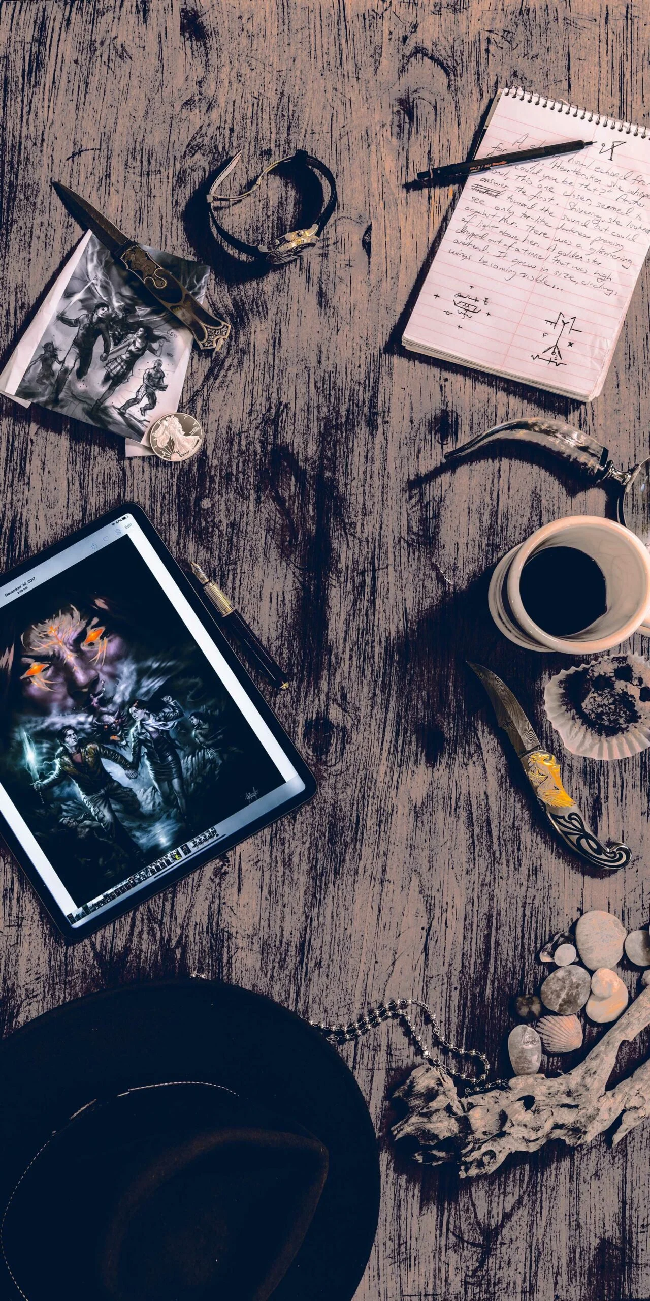 A wooden table with various items, including a digital tablet displaying digital art, a notebook with handwritten notes, a pen, a cup of coffee, a decorative knife, a skull necklace, rocks, seashells, and a black hat.