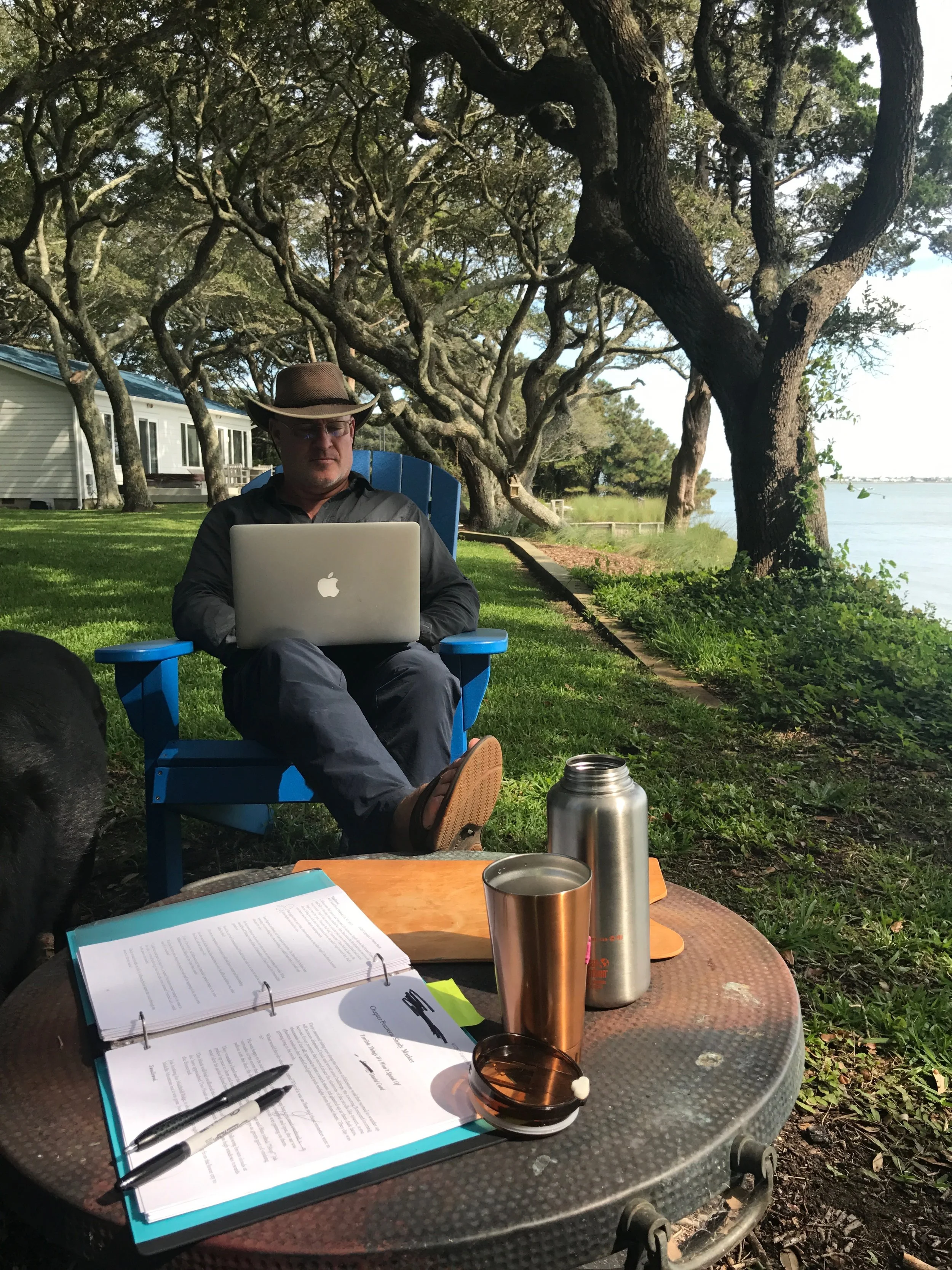 Man sitting outdoors at a table with documents, pens, and drink containers, surrounded by trees and a body of water in the background.