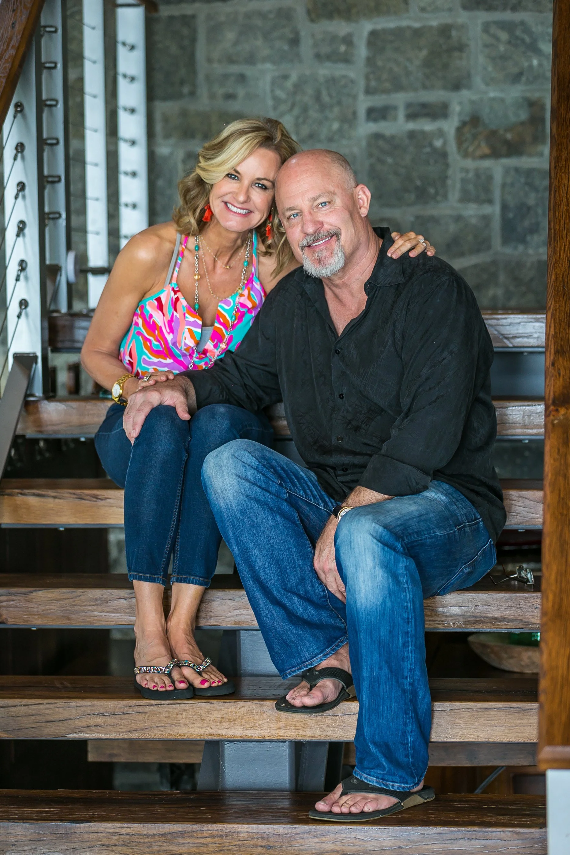 A smiling woman and man sitting on wooden stairs indoors, with a stone wall background, close together, with the woman resting her hand on the man's shoulder.