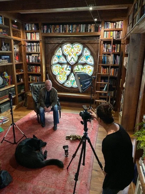 A man sitting on an armchair in a cozy library room with wooden bookshelves filled with books. Behind him is a decorative stained glass window with a floral design. A camera crew, including a woman operating a camera on a tripod and a black dog lying on a red rug, is filming the man.
