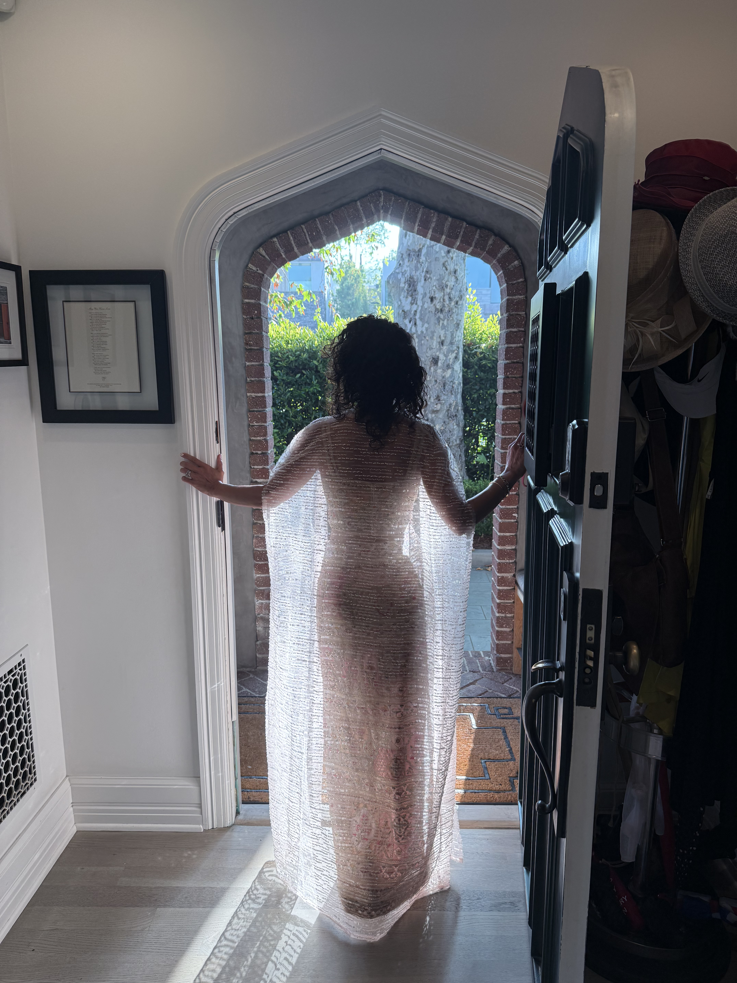 A woman standing in an open doorway, wearing a translucent, beaded, floor-length dress, with sunlight illuminating her from behind, revealing her silhouette.