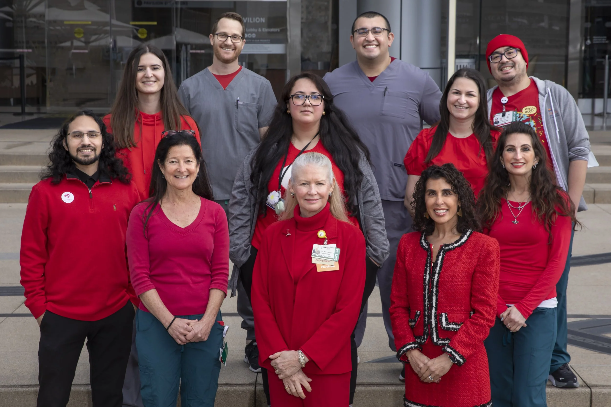 Group of healthcare professionals standing outside a building, smiling, wearing scrubs, name tags, and red clothing, with stairs in the background.