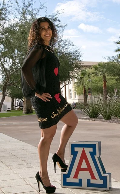 Young woman with curly hair posing outdoors in front of a University of Arizona sign, wearing a black dress with red heart designs and black high heels.