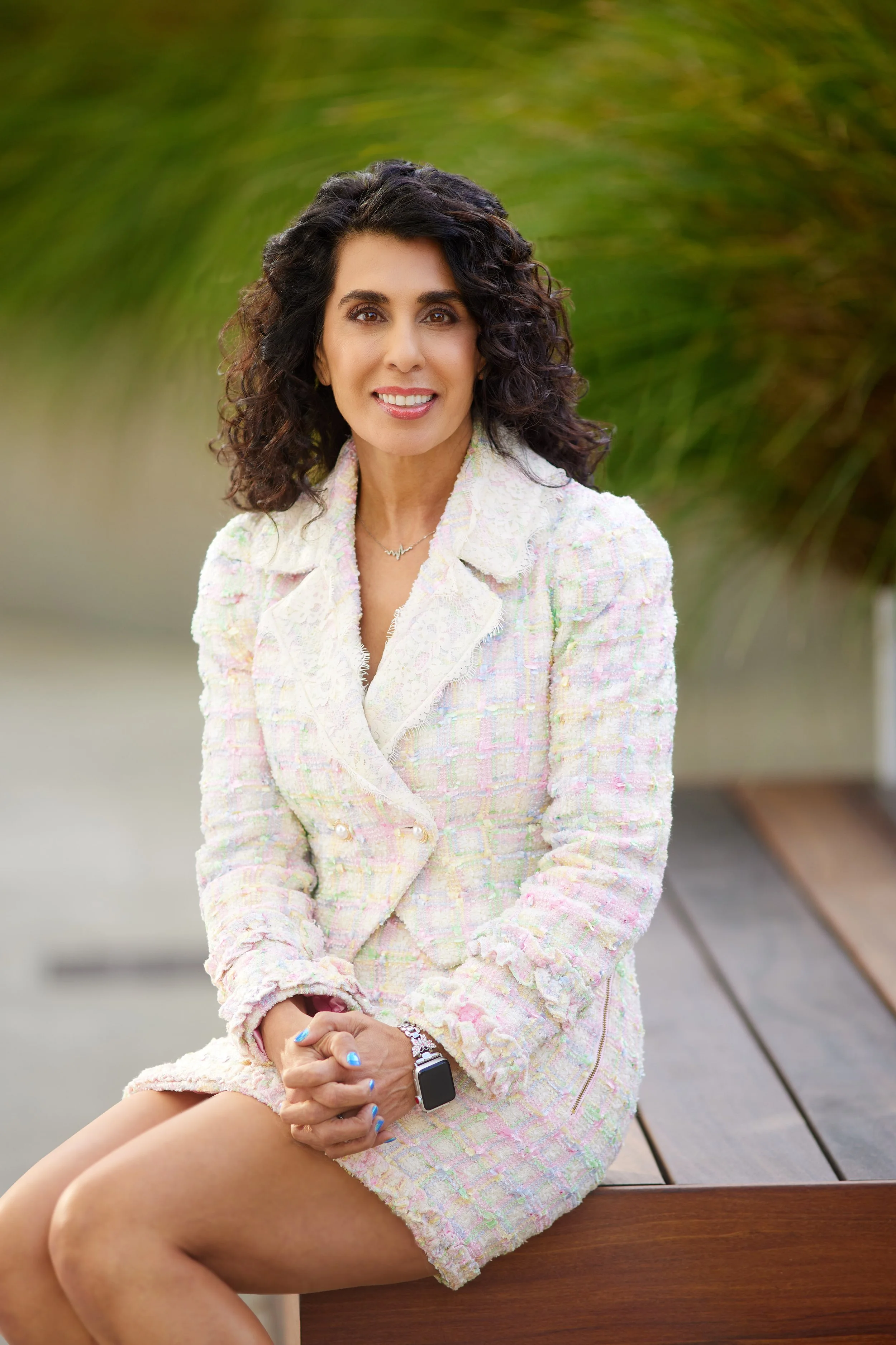 A woman with curly dark hair, wearing a pastel tweed blazer, sitting on a wooden bench outdoors with blurred green foliage in the background.