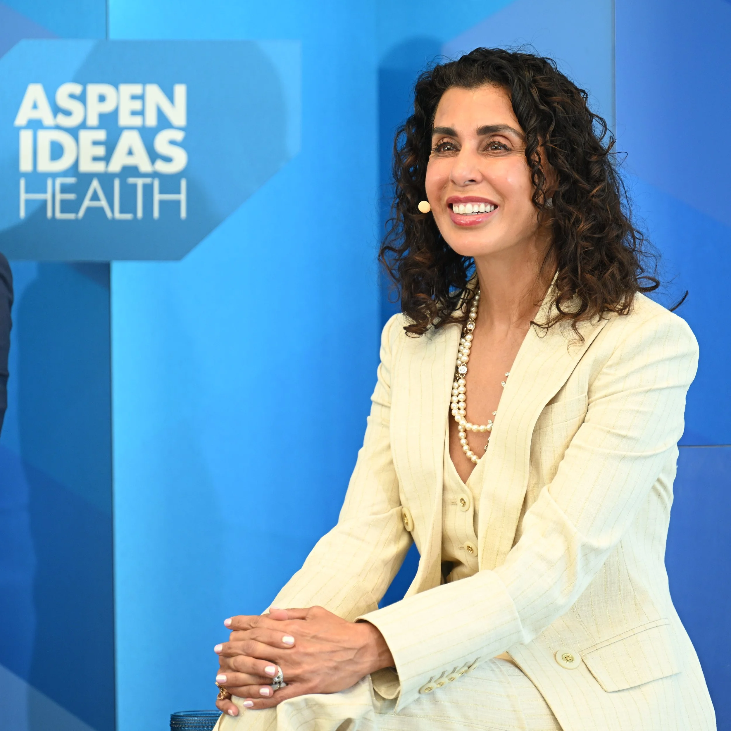A smiling woman with curly dark hair, wearing a light-colored blazer and pearl jewelry, seated with her hands clasped together, at an event with a blue backdrop that reads 'Aspen Ideas Health'.