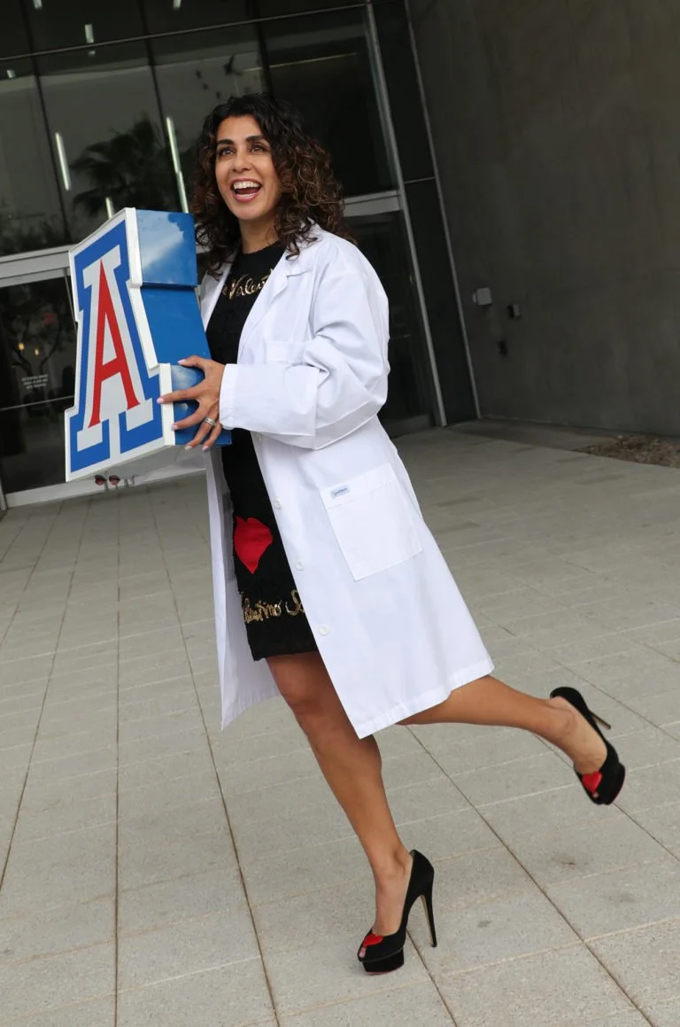 A woman in a white coat holding an Arizona Wildcats box outside a building, smiling and standing on one leg with the other leg raised behind her.