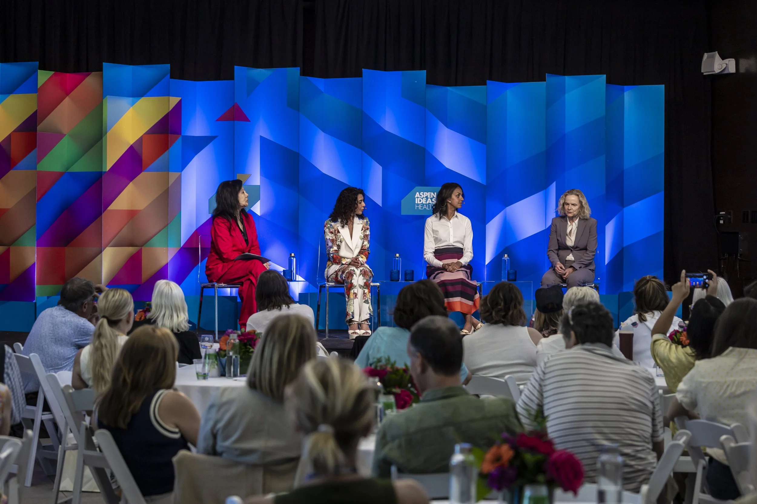 A panel of four women speaking at an event with an audience seated at tables and taking photos. The backdrop is a colorful, geometric-patterned blue screen with the sign "Aspen Ideas Health".