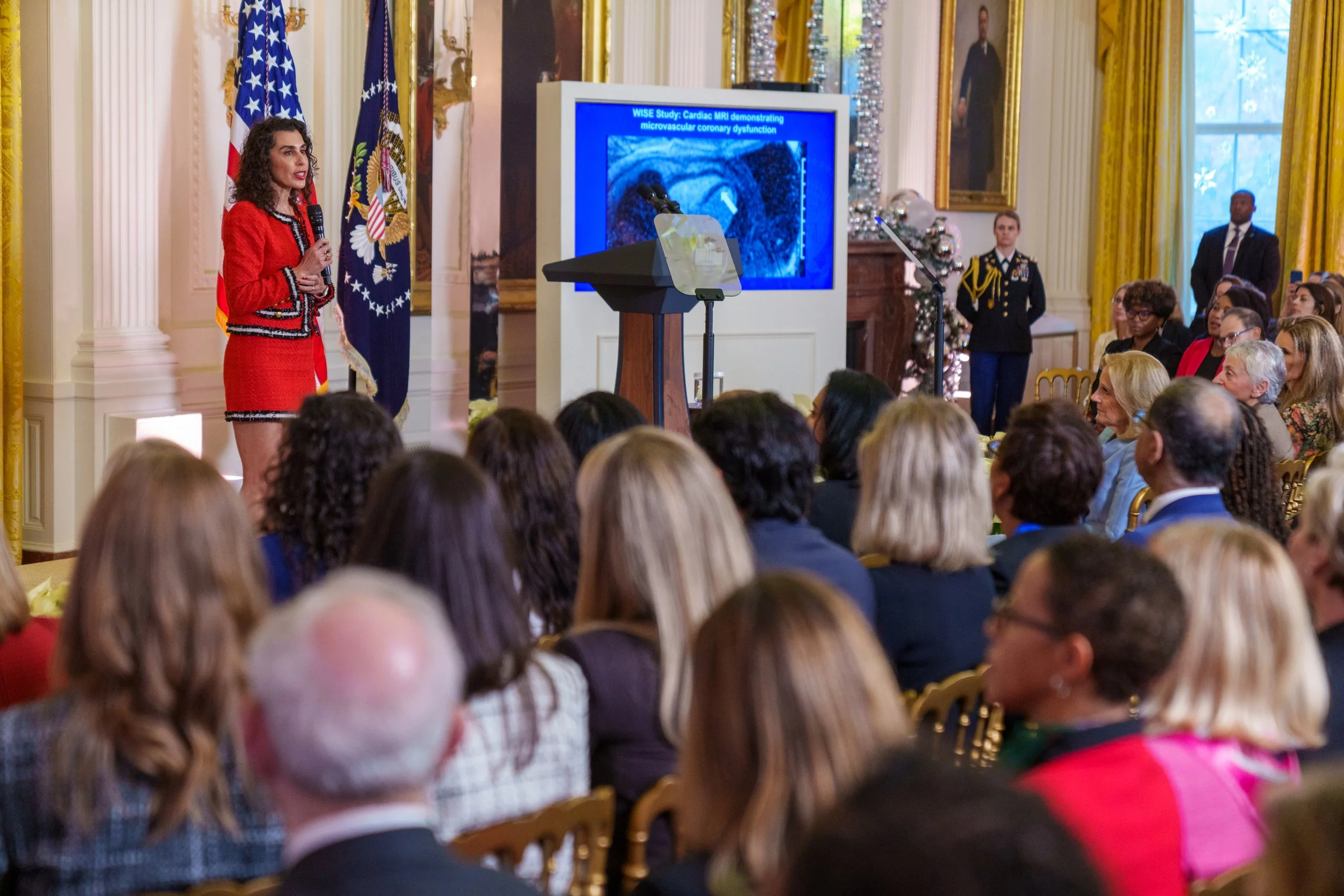 A woman in a red jacket and black skirt speaking into a microphone at a presentation in a decorated room with American flags and a large screen displaying medical imagery.