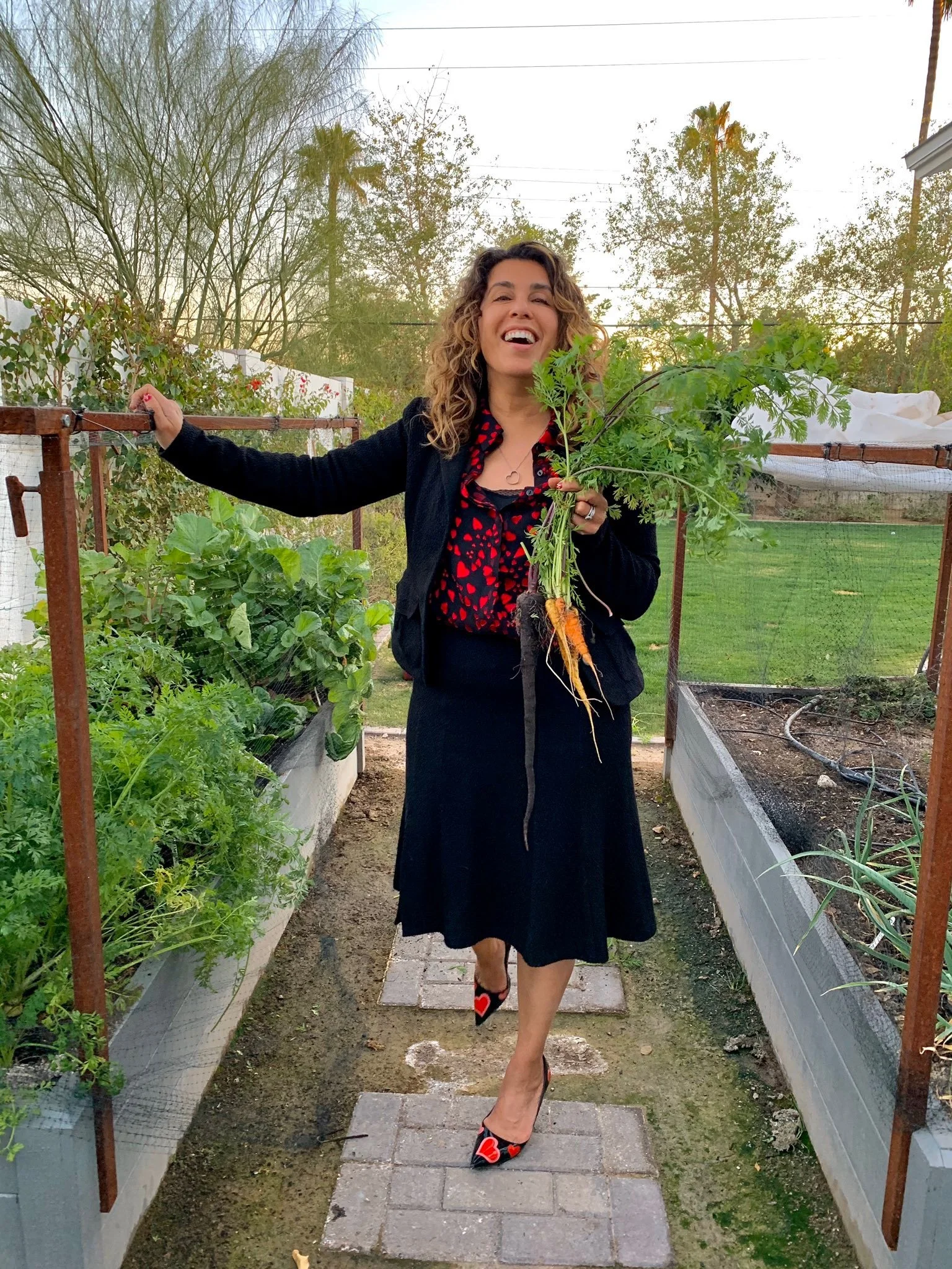 Woman holding a bunch of freshly picked carrots and smiling while standing on a garden path surrounded by vegetable garden beds.