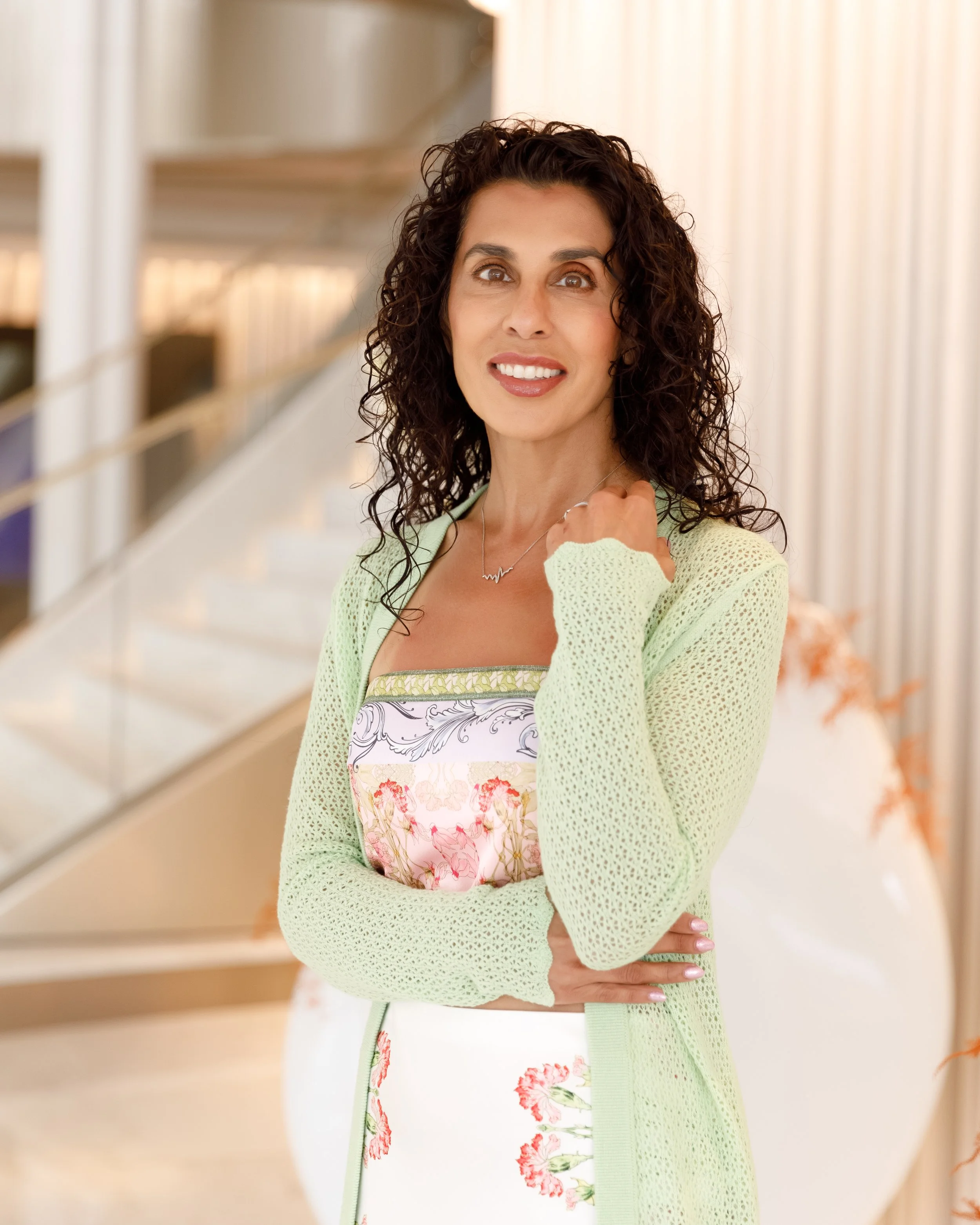A woman with curly dark hair standing indoors, wearing a pastel-colored dress with floral patterns and a light green knit cardigan, smiling and looking at the camera.