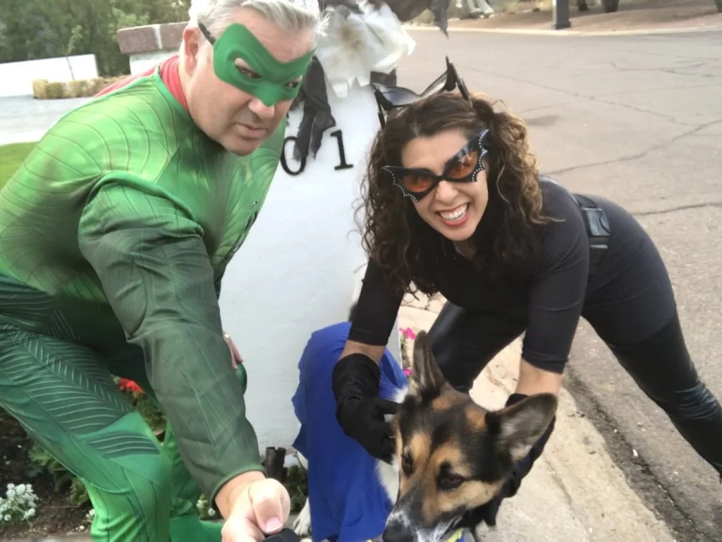 A person dressed as the Green Goblin from Spider-Man with a green costume and mask, and a woman wearing sunglasses with bat-shaped frames, both smiling while petting a dog. The woman is crouched near the curb, and the dog is wearing a blue coat.