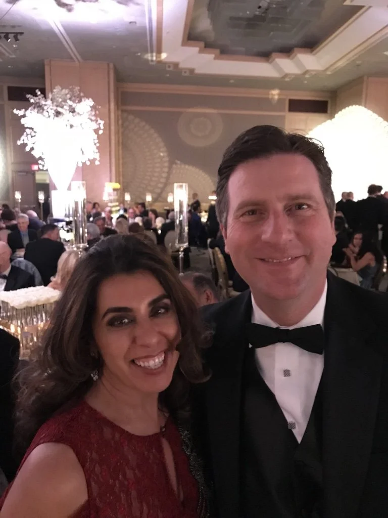 A smiling woman in a red dress and a man in a tuxedo taking a selfie at an elegant formal event in a decorated banquet hall filled with guests, floral arrangements, and ambient lighting.