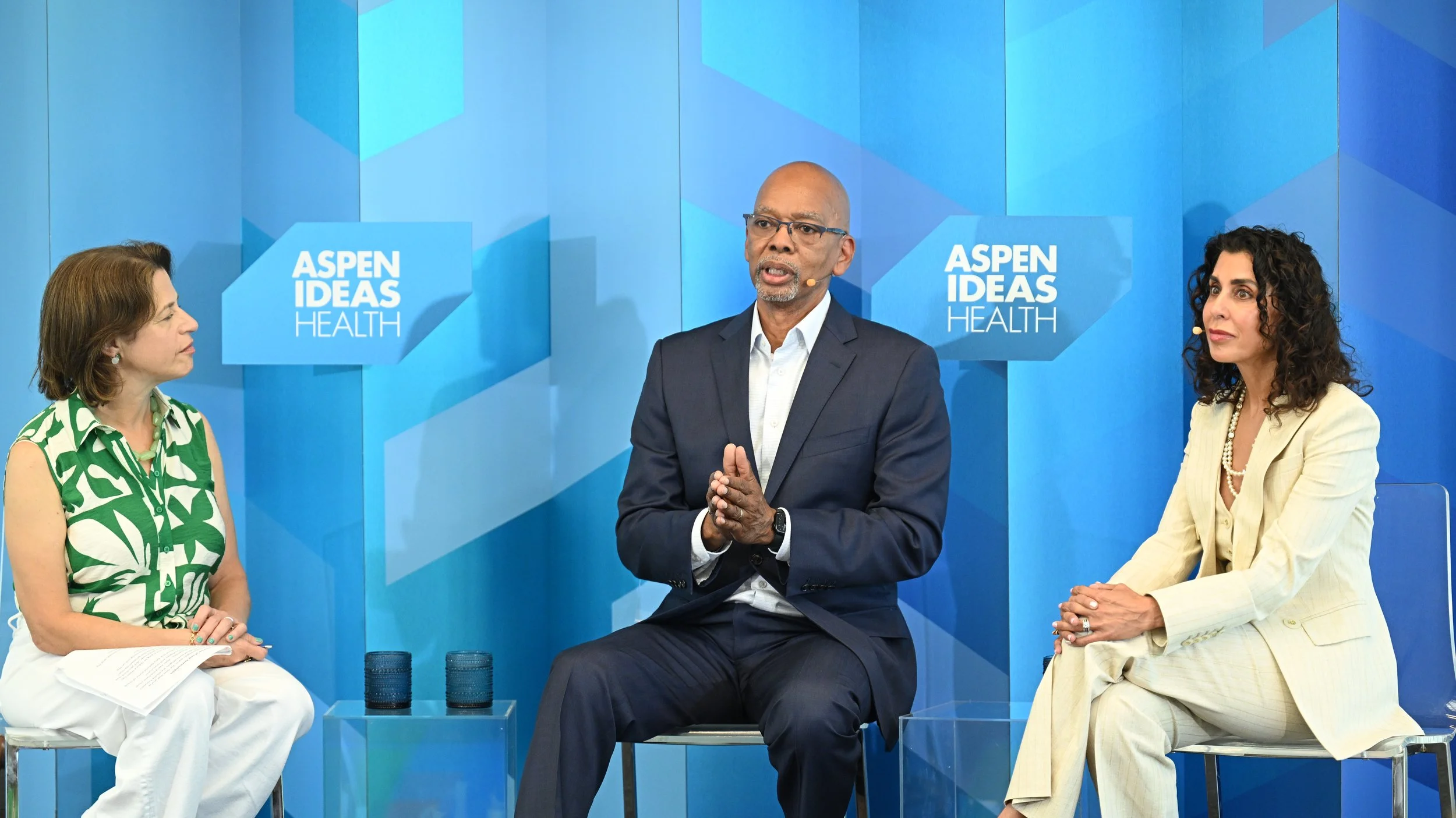 Three people sitting on stage during a panel discussion at Aspen Ideas: Health, with a blue geometric background and the event logo.