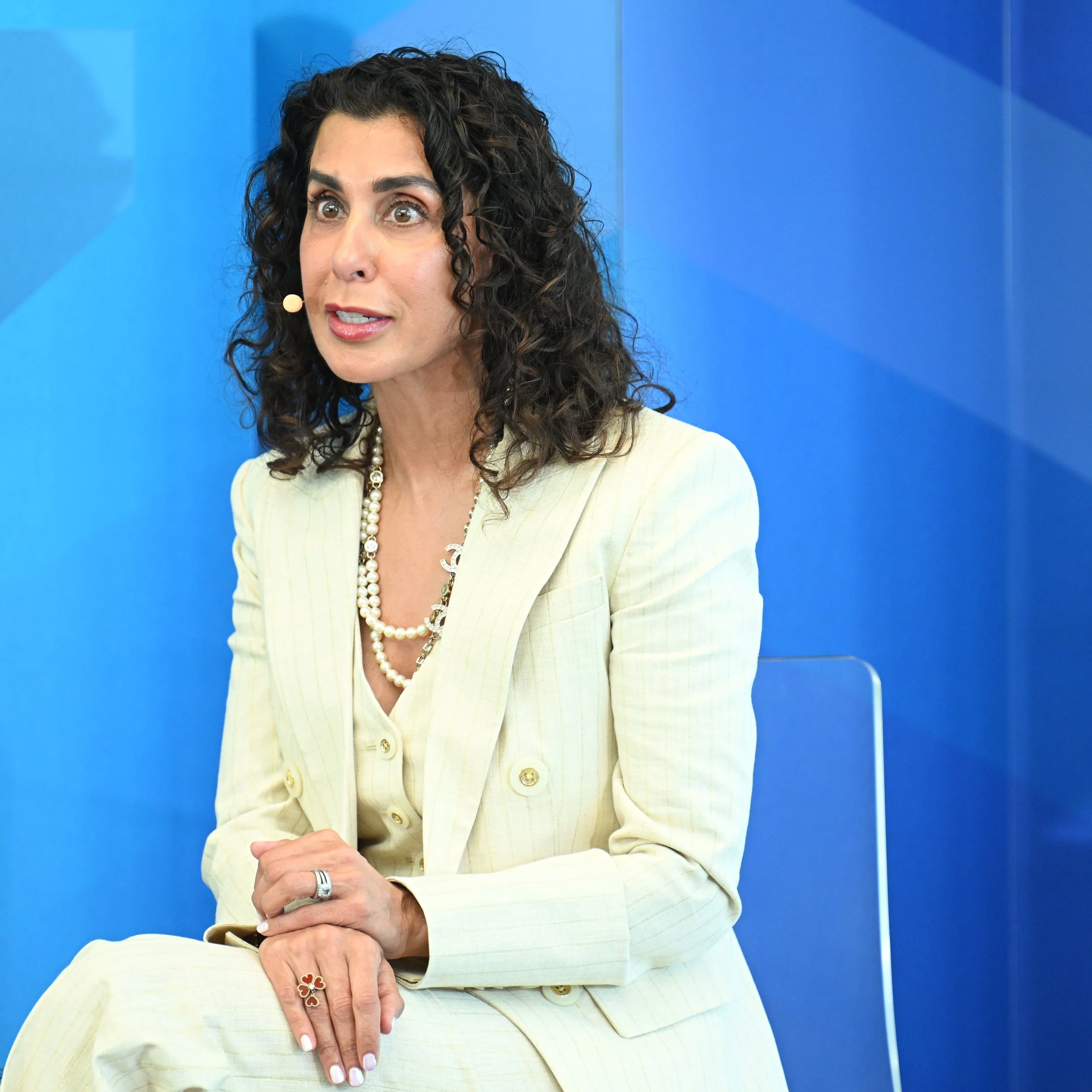 A woman with curly dark hair, wearing a light yellow suit and pearl jewelry, sitting against a blue background.