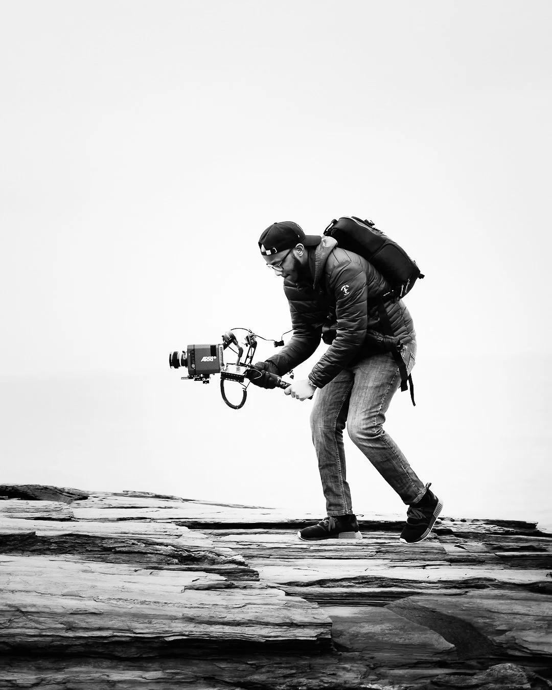 Black and white photo of a man filming with a professional camera on a tripod outdoors, standing on rocky terrain.