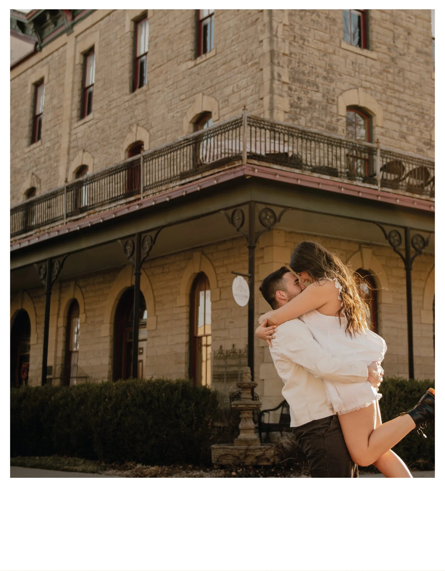 A man lifting a woman in front of a historic brick building with iron balconies, during sunset
