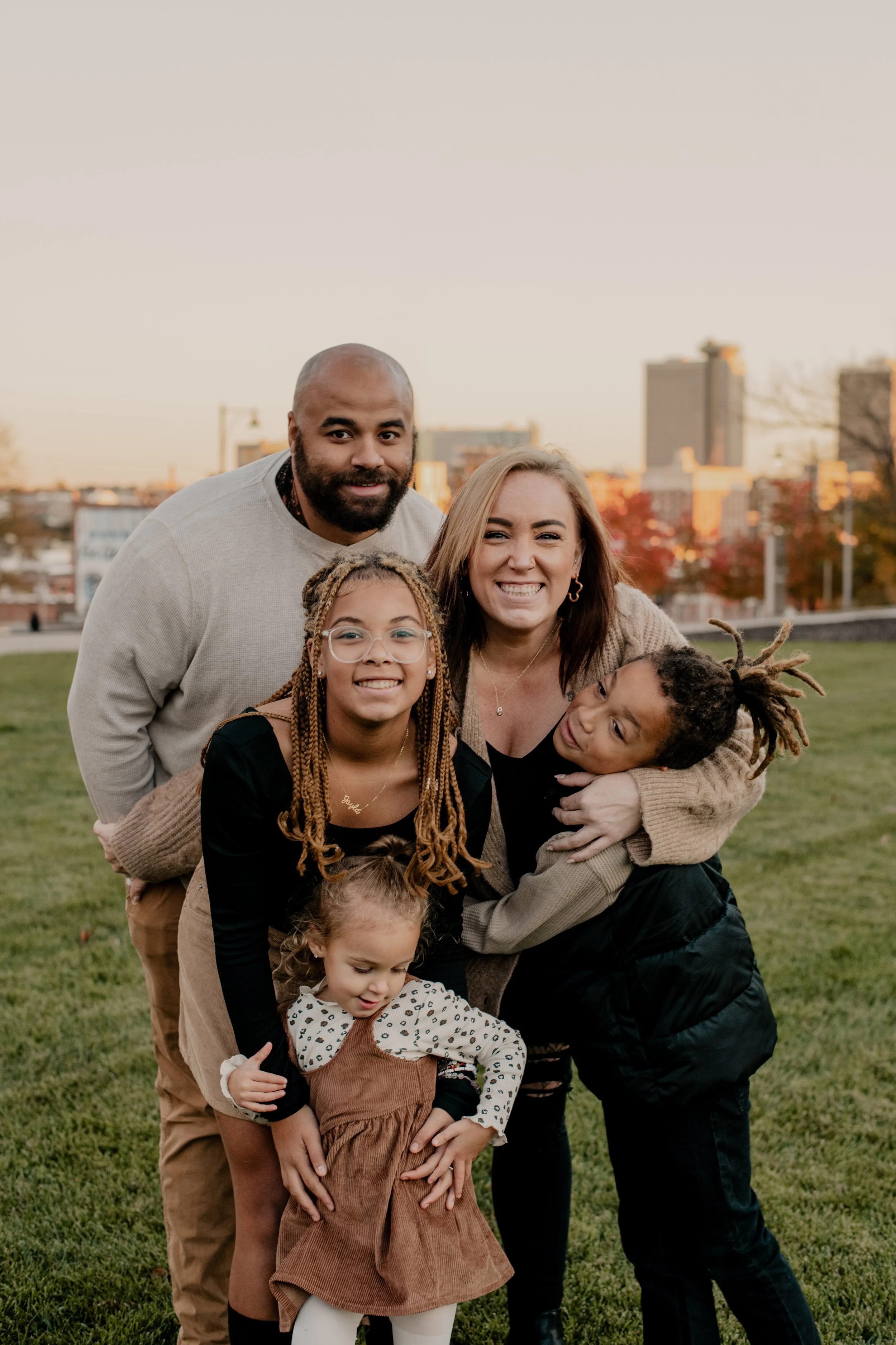 A family six people, including children and adults, smiling and posing together outdoors on a grassy field during sunset, with city buildings in the background outside the Kauffman Performing Arts Center in Kansas City, Missouri.