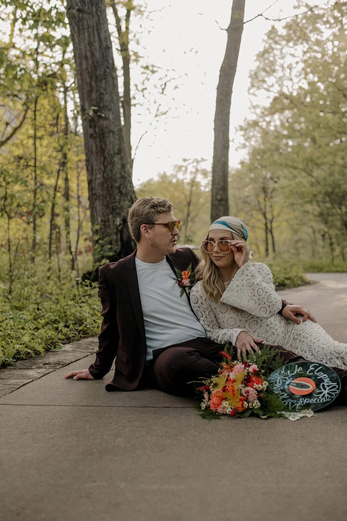 A man and woman sit on a paved path in a wooded area, dressed in vintage-style clothing and wearing sunglasses, surrounded by flowers, with a handmade sign that reads, "We Eloped" at the Powell Gardens in Kansas City, Missouri
