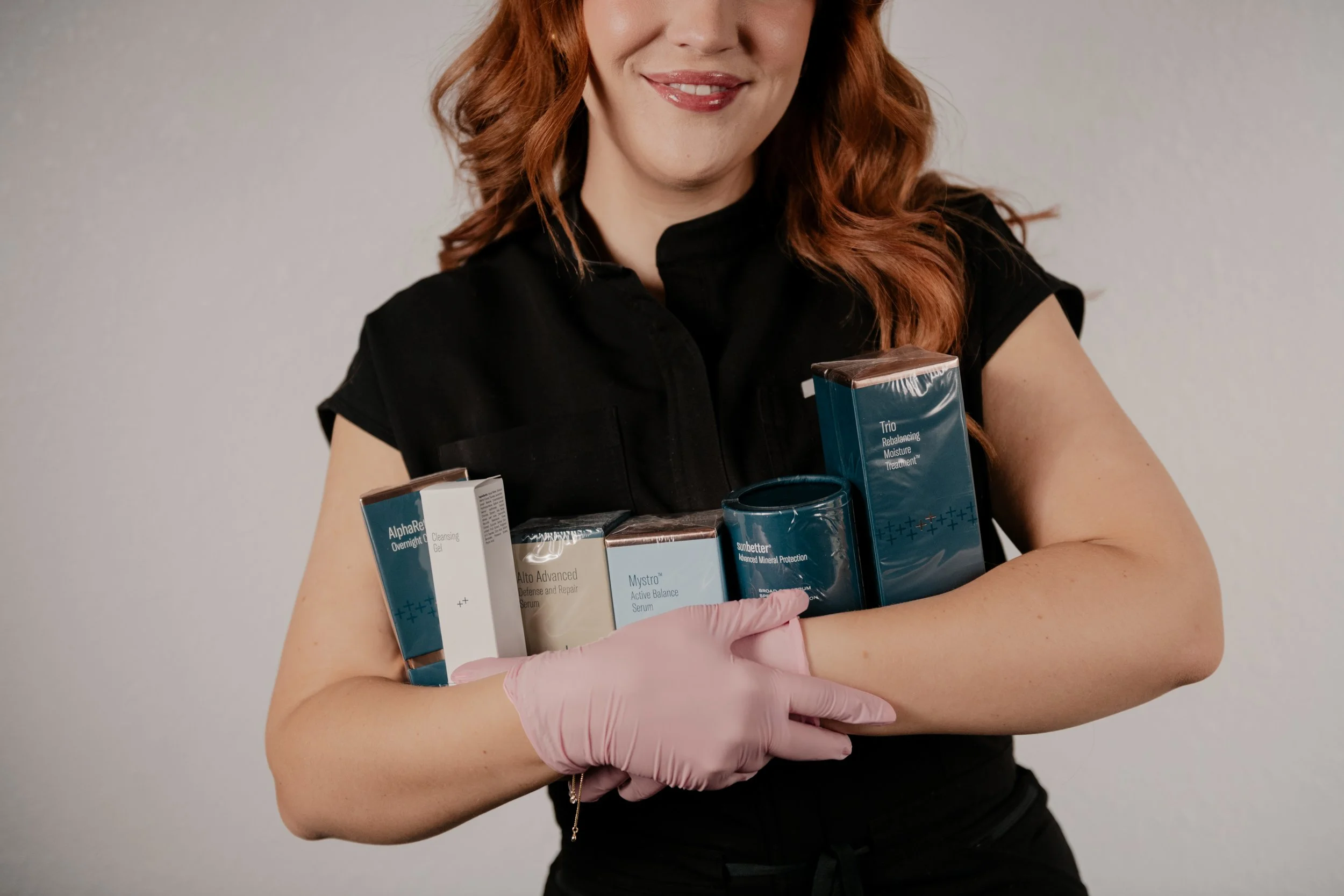 A red headed woman with pink gloves holding six skin care products in her arms.