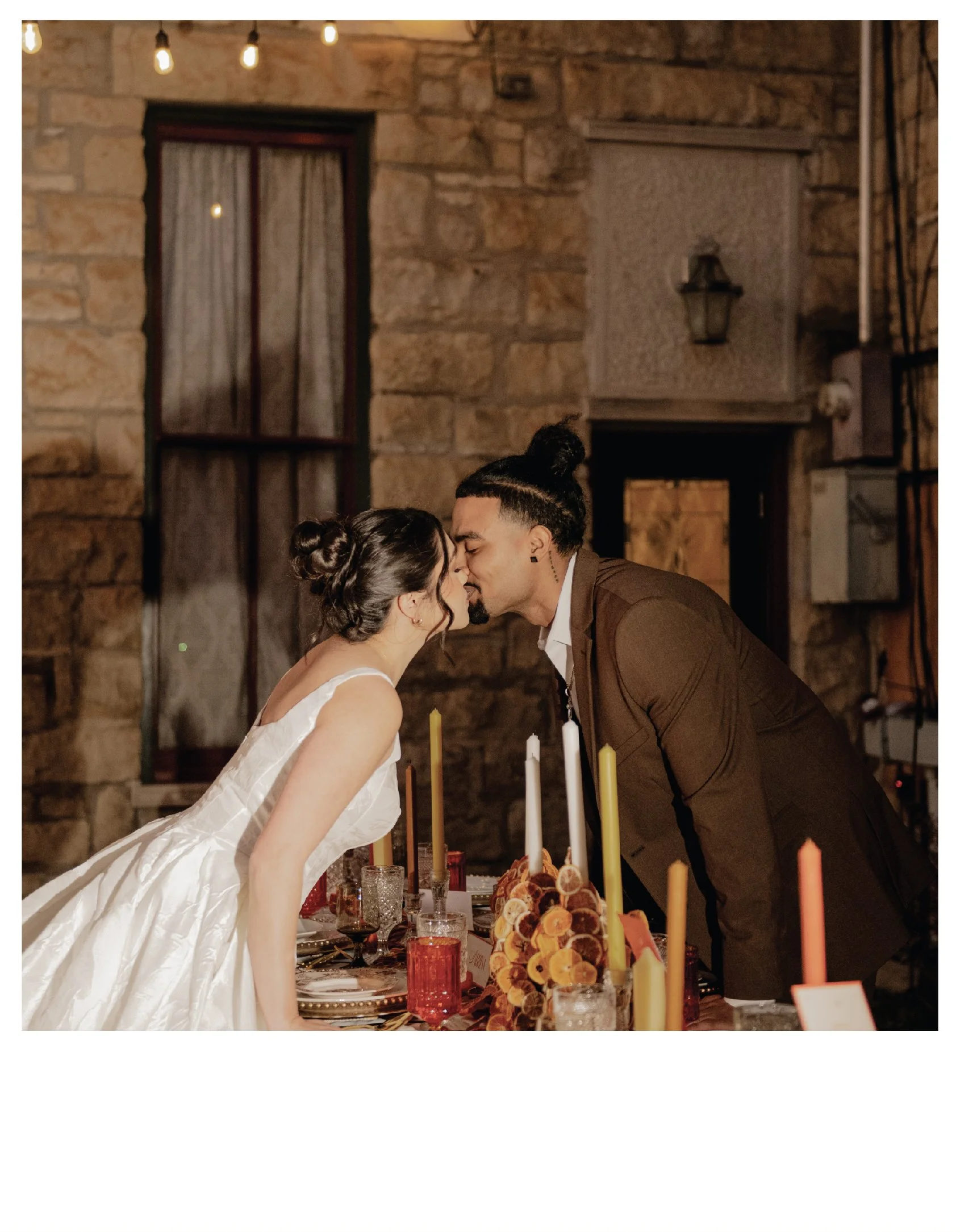 Couple at wedding, leaning in for kiss, at decorated reception table with candles and floral centerpiece, in rustic indoor setting with stone walls. at the Historic Elgin Hotel in Marion, Kansas.