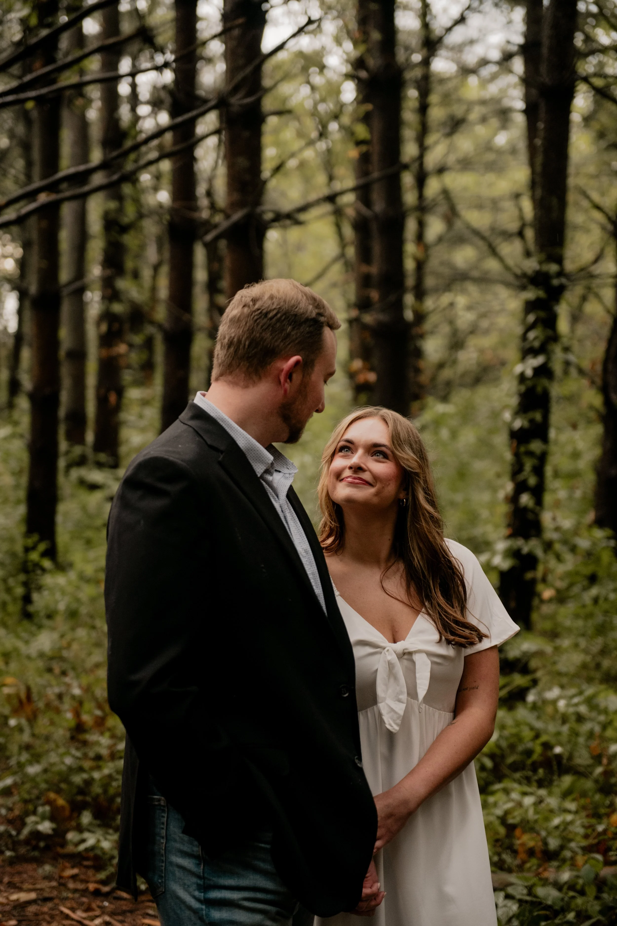  A pictured of a young couple in the mid 20’s standing in the middle of a forest at the Burr Oaks Conservation Area posing for an engagement portrait in the Kansas City Metro. The woman is staring lovingly at the man. 