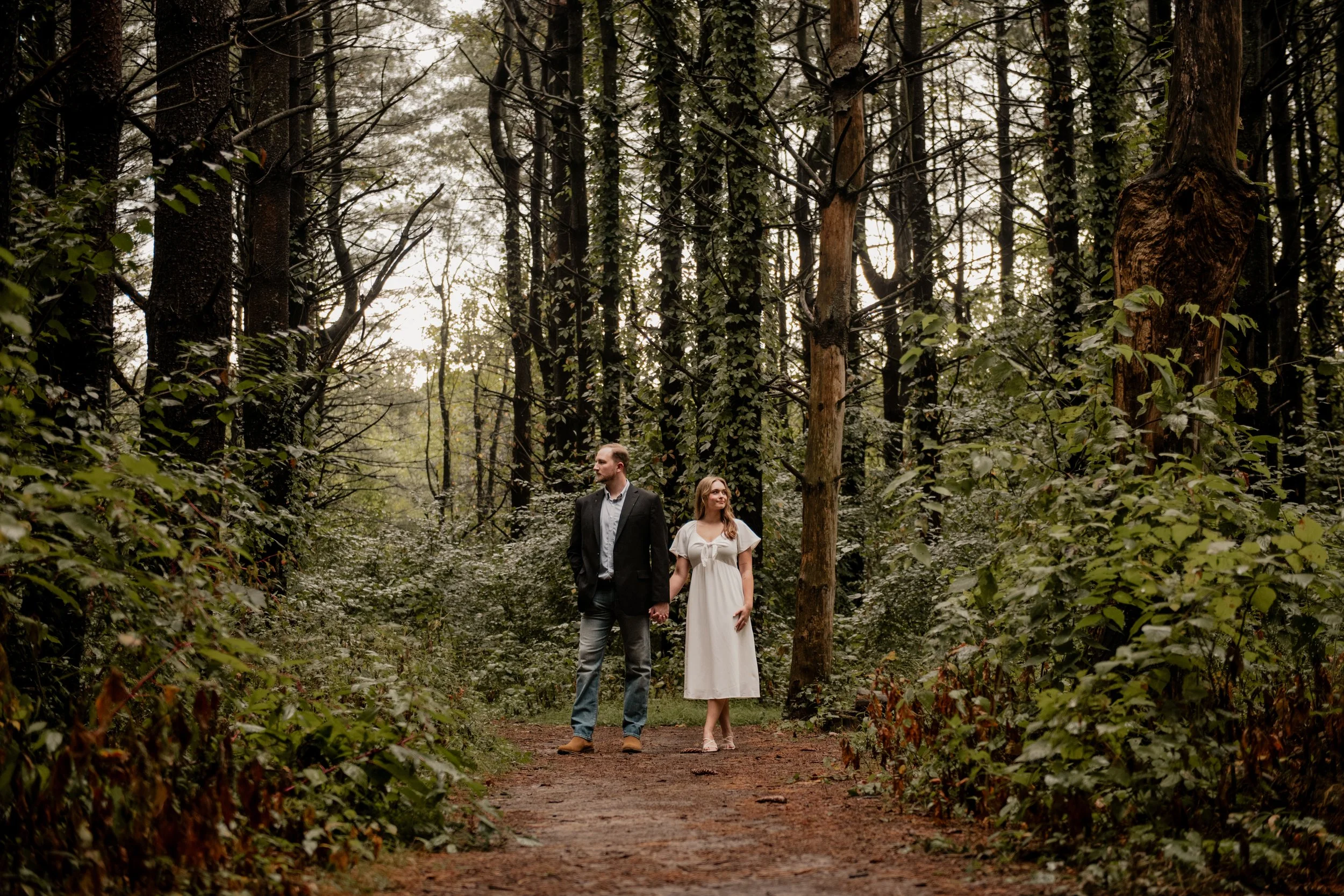  A pictured of a young couple in the mid 20’s standing in the middle of a forest at the Burr Oaks Conservation Area posing for an engagement portrait in the Kansas City Metro. The couple stand next to each other hand in hand while they look in opposi